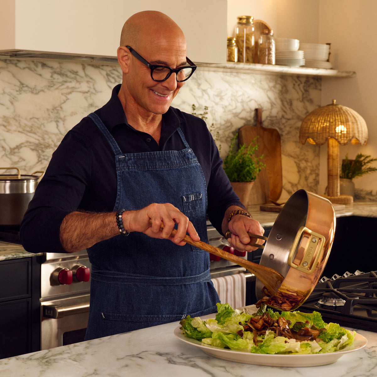 Person in apron serves food from a copper Stanley Tucci 4-quart sauté pan onto salad in a modern kitchen.