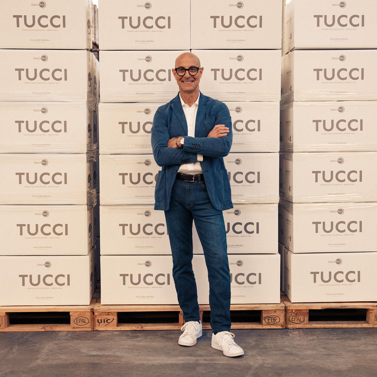 Man in blue jeans and blazer stands smiling with arms crossed next to Stanley Tucci Copperply sauté pan boxes.