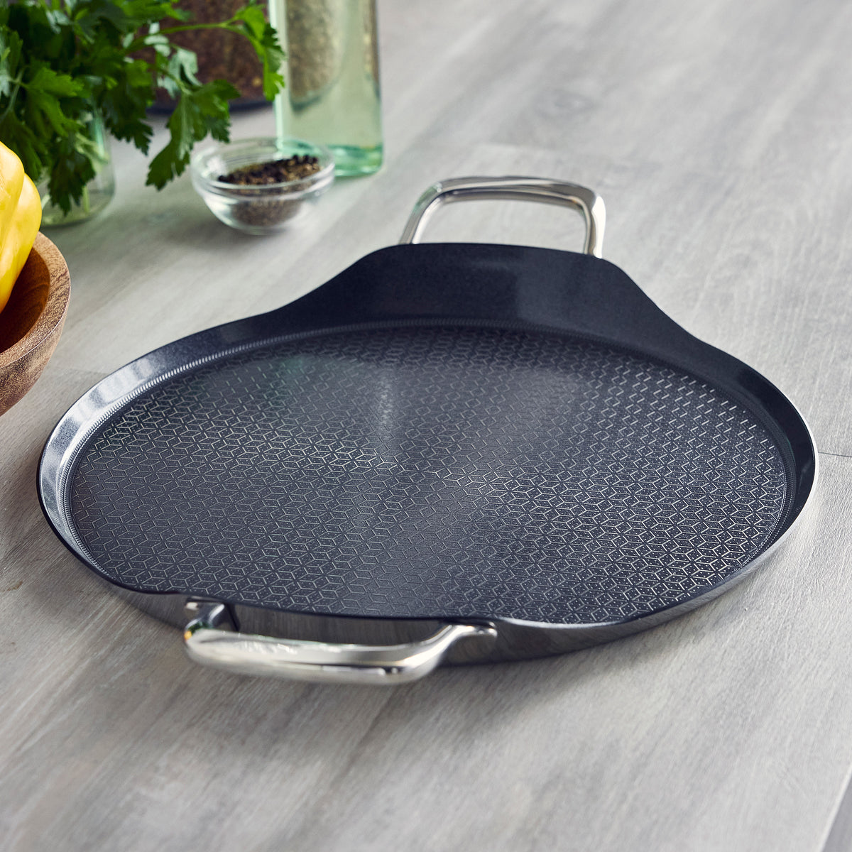 Black 13-inch round Spectra Thermobond hybrid ceramic griddle with two stainless steel handles and a textured hexagonal surface.