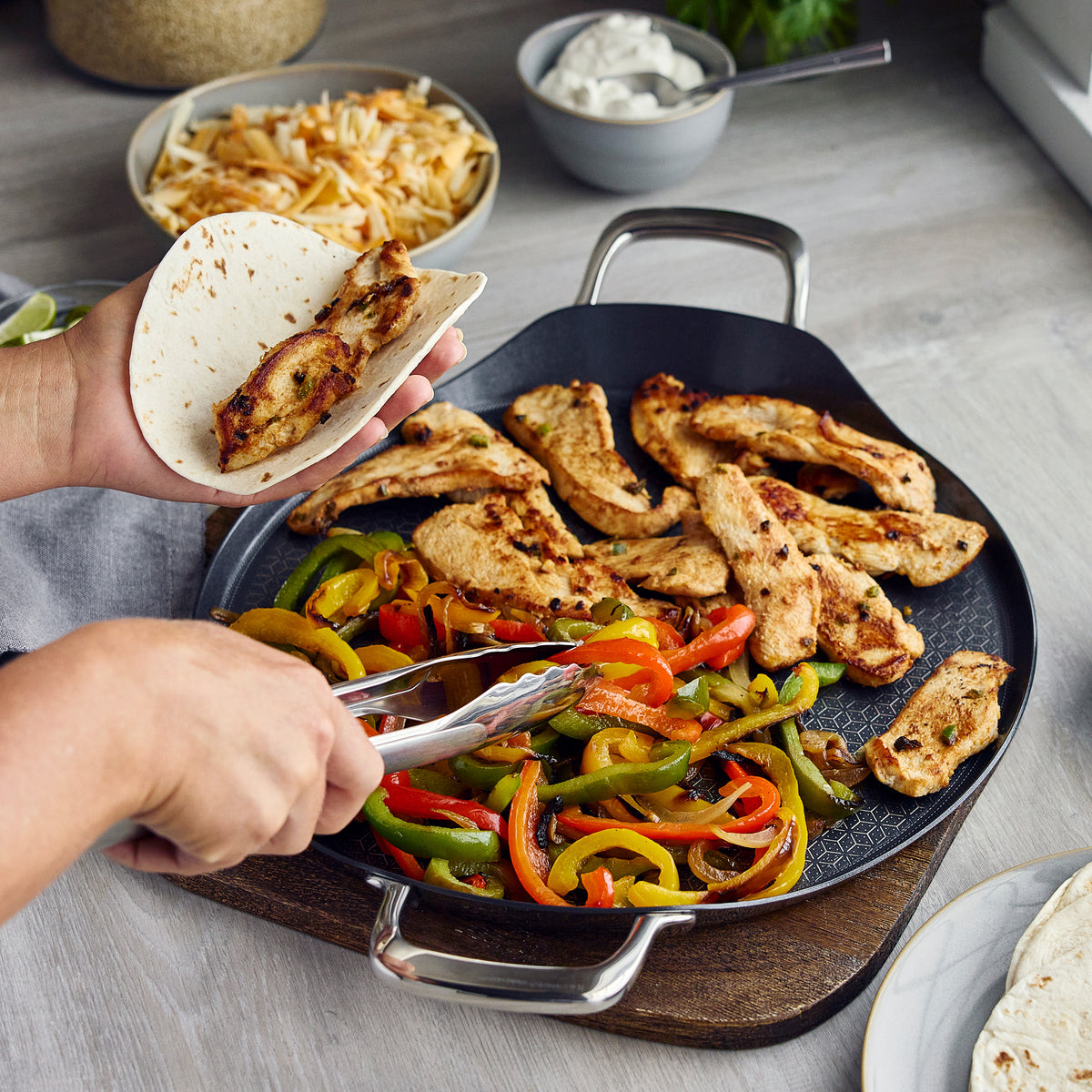 Spectra Thermobond hybrid ceramic 13″ round griddle cooking chicken and peppers with tongs and a tortilla.