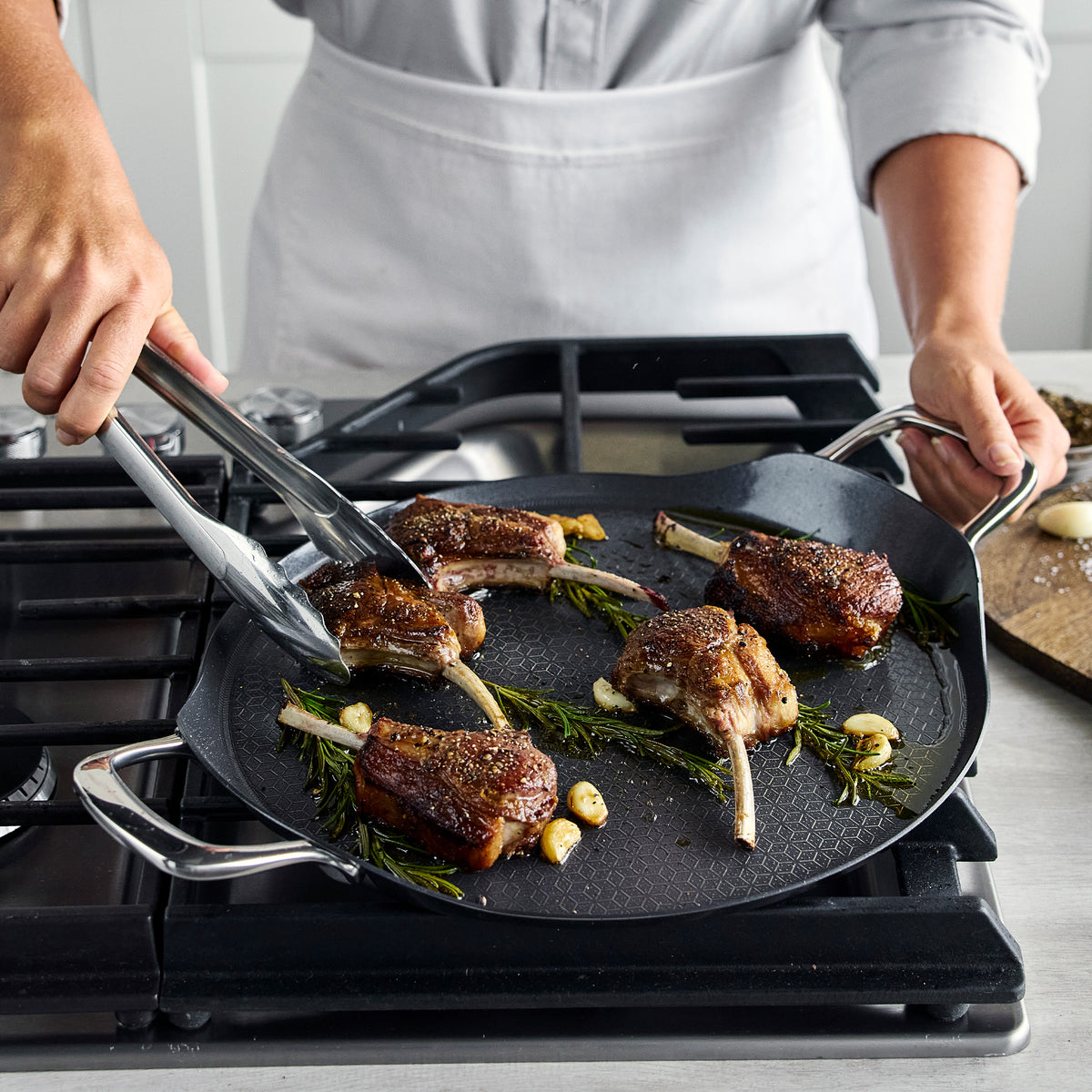 Metal tongs turning lamb chops on a 13″ Spectra Thermobond hybrid ceramic nonstick griddle.