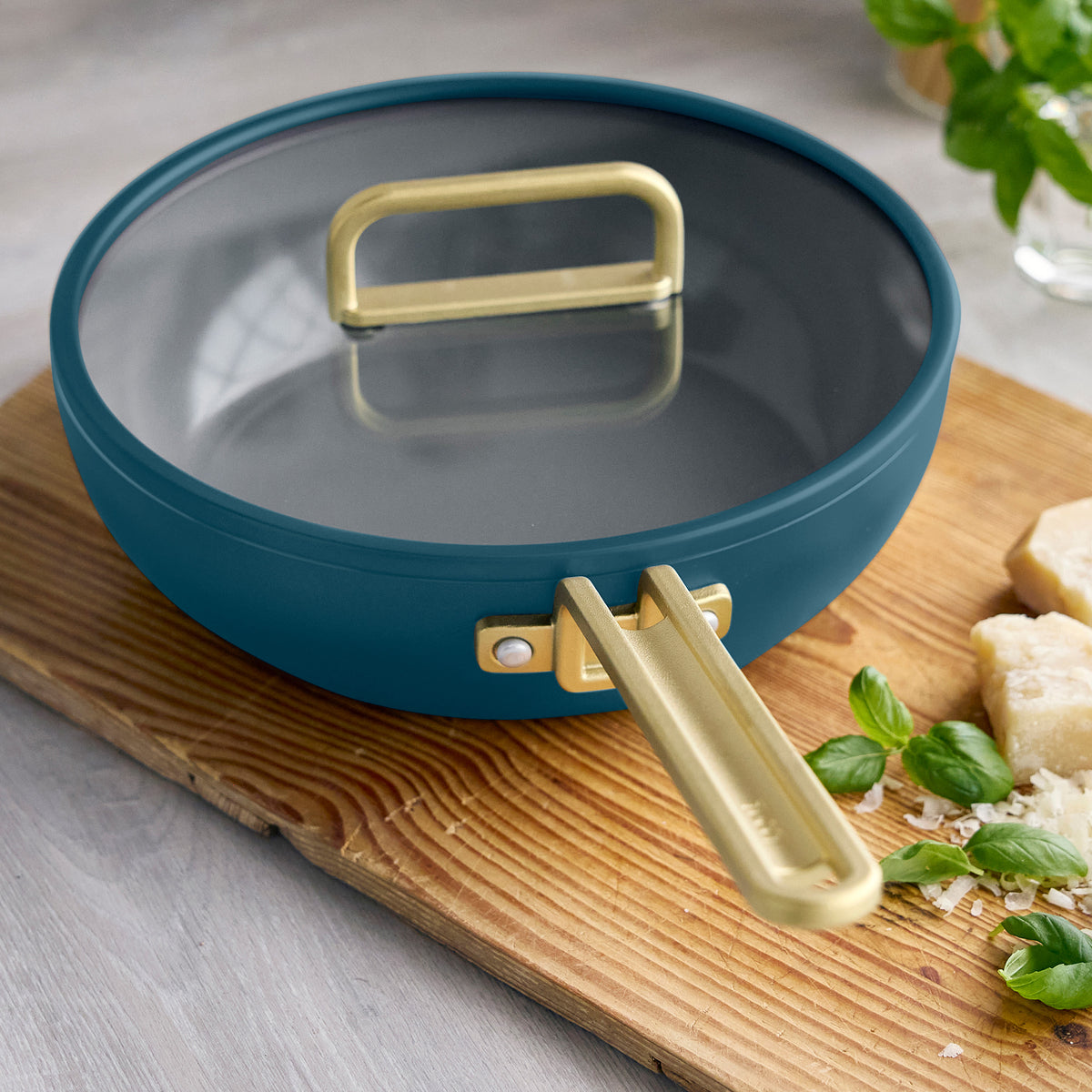 Stanley Tucci 3.25-quart ceramic nonstick piccolo pan in Venetian Teal with a gold handle and glass lid, displayed with cheese and basil.
