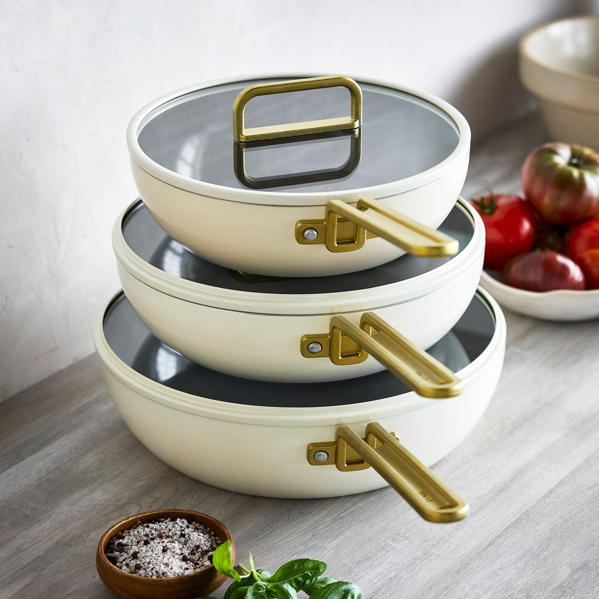 Stack of three Carrara White Stanley Tucci 3.25-quart ceramic nonstick pans with gold-toned handles and glass lids.
