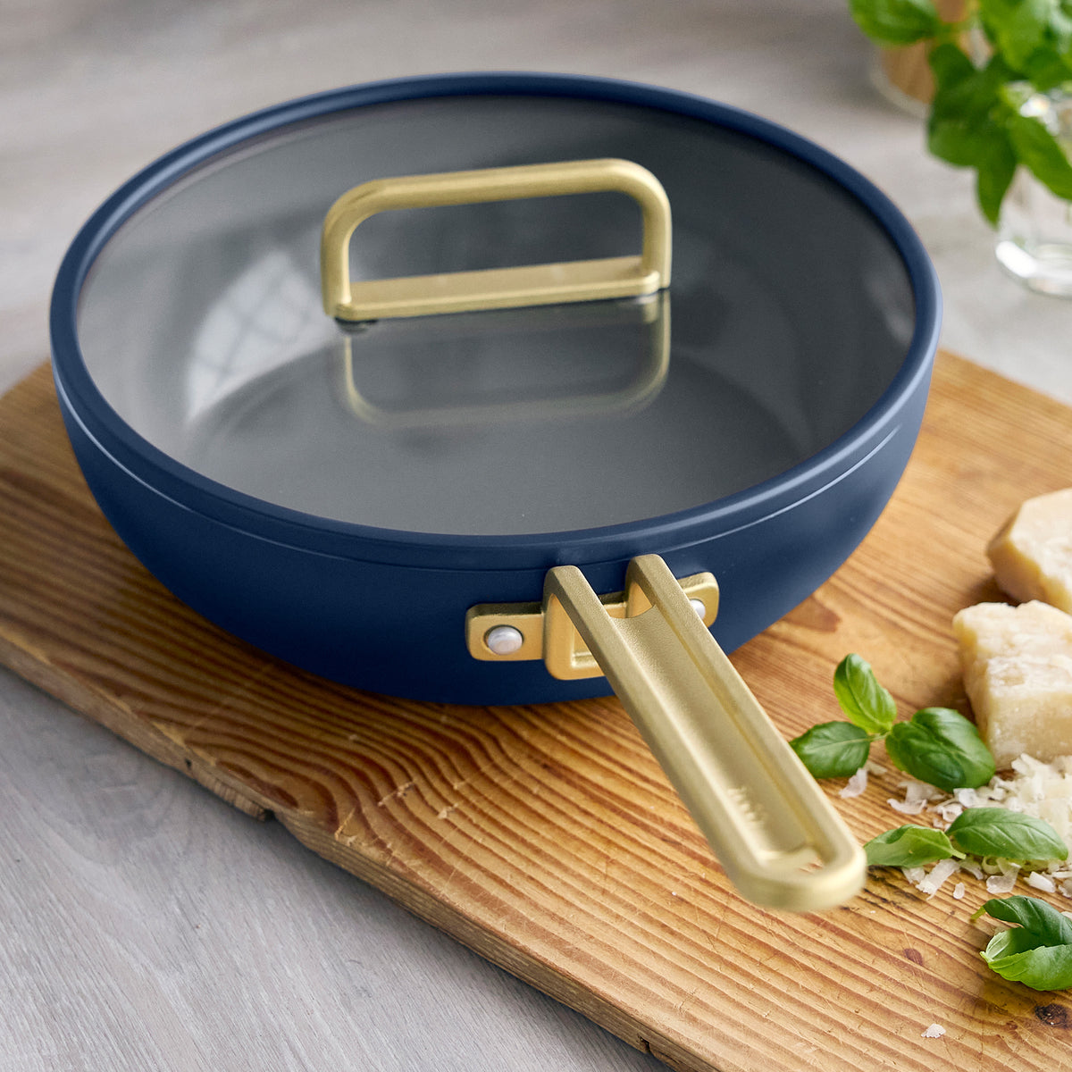 Stanley Tucci 3.25-quart ceramic nonstick Piccolo pan in Marino Blue with a glass lid and gold-tone handle on a wooden cutting board.