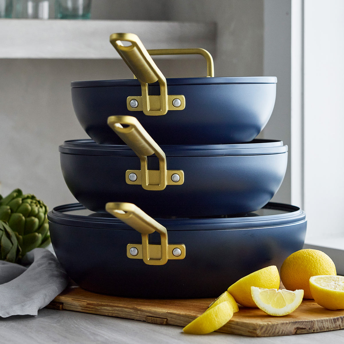 Three stacked 3.25qt Marino Blue Stanley Tucci™ Piccolo ceramic nonstick pans with gold handles on a cutting board with lemons.