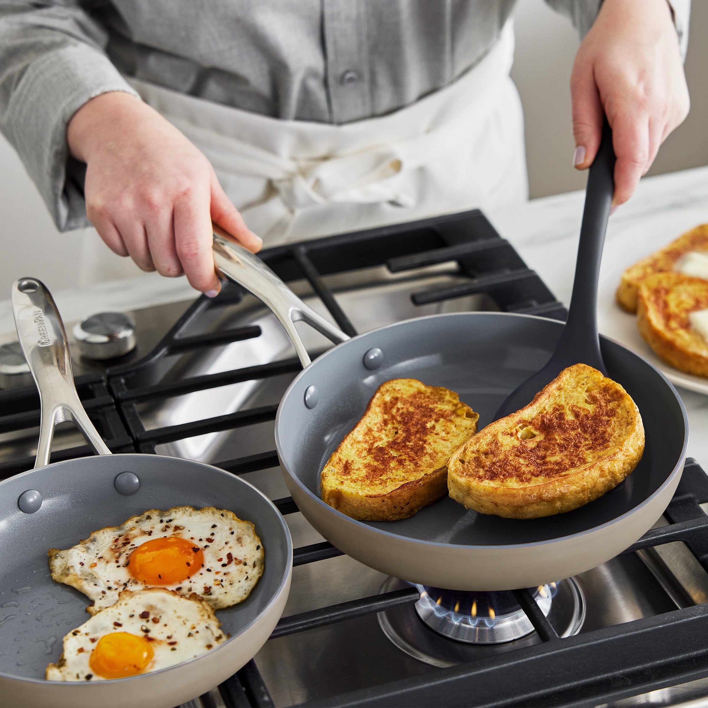 Taupe ceramic nonstick frypan set with eggs and French toast cooking on a stovetop, two pans visible.