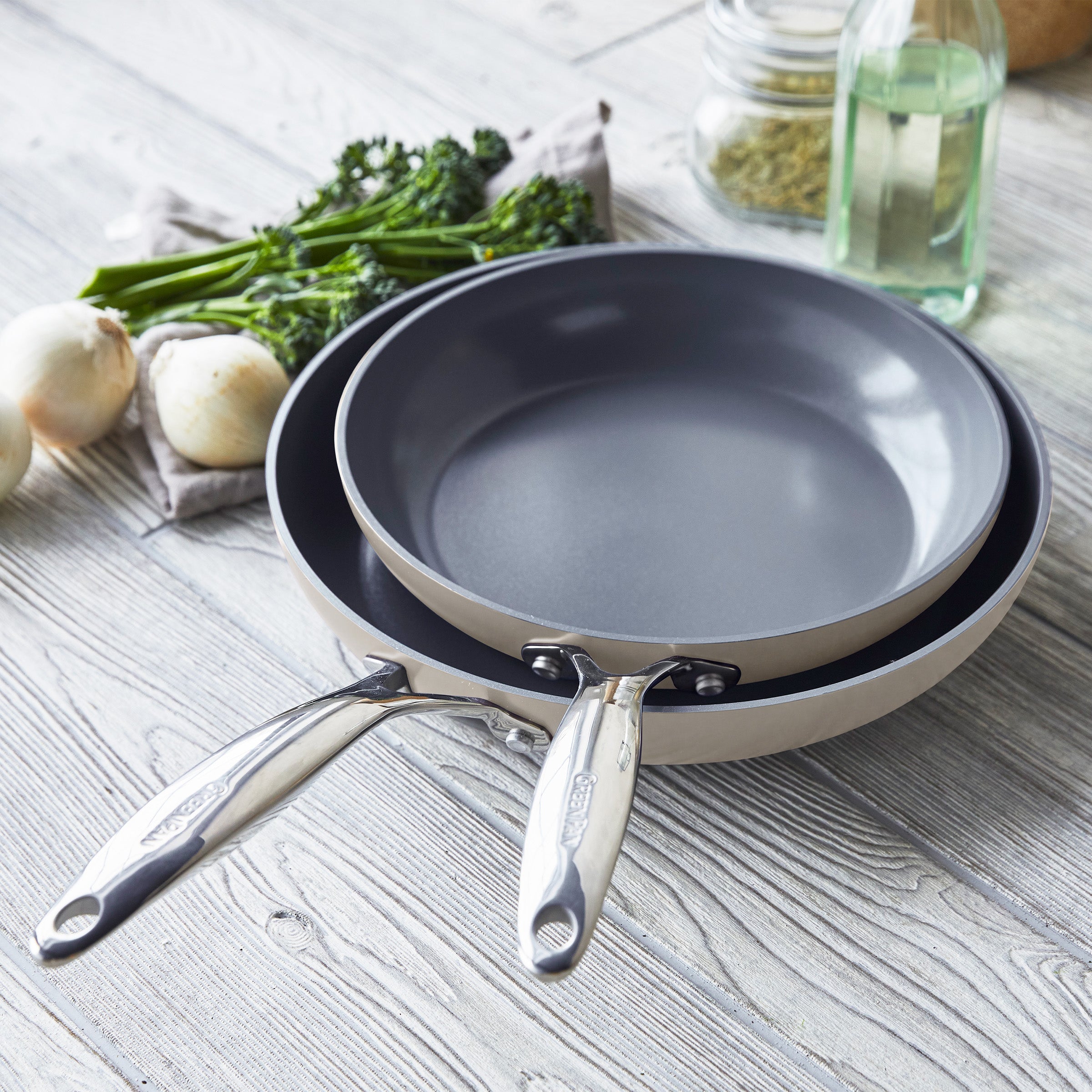 Two taupe ceramic nonstick frypans with metal handles next to onions, broccolini, and a bottle of oil.