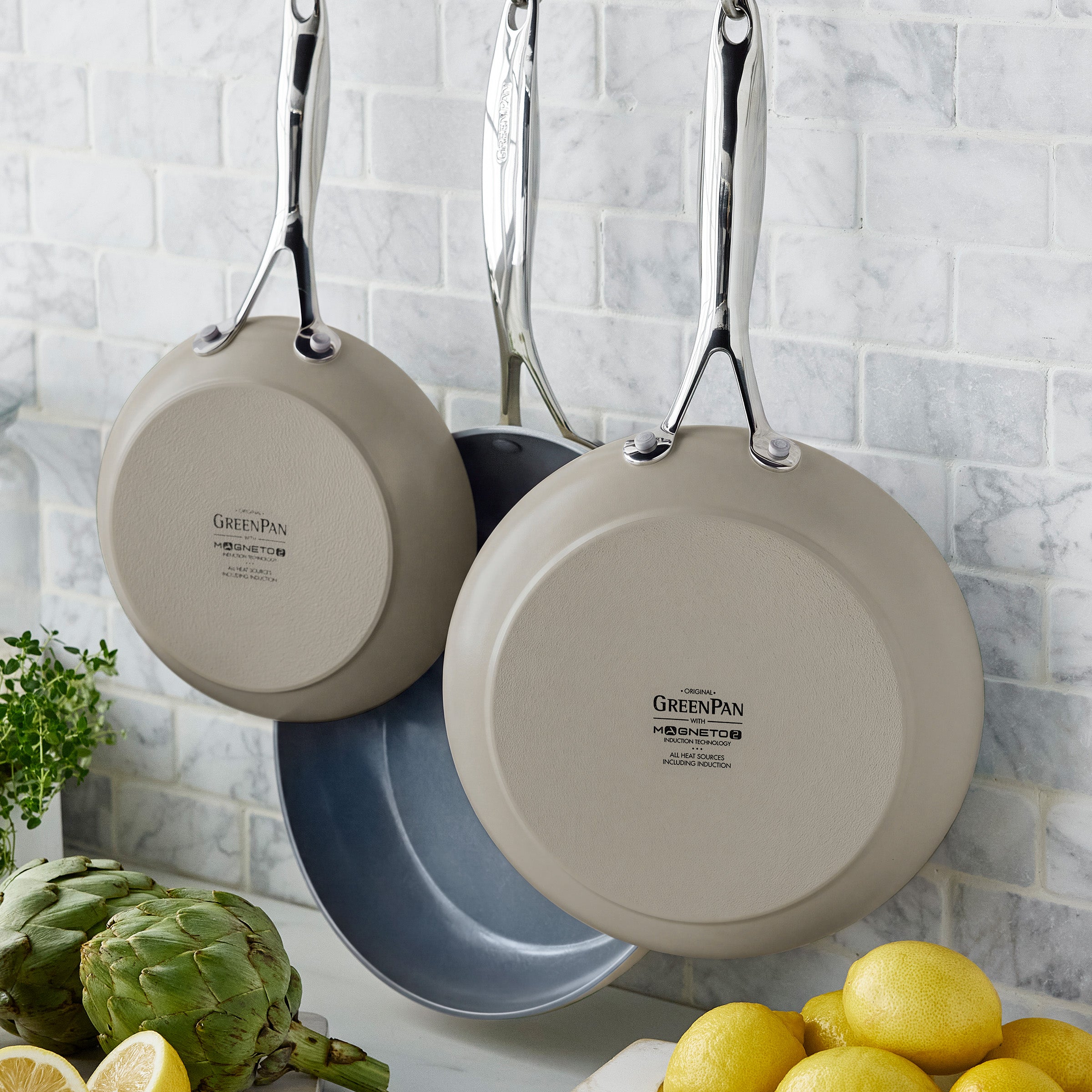 Three taupe nonstick frypans hanging above a marble counter with lemons and assorted vegetables.