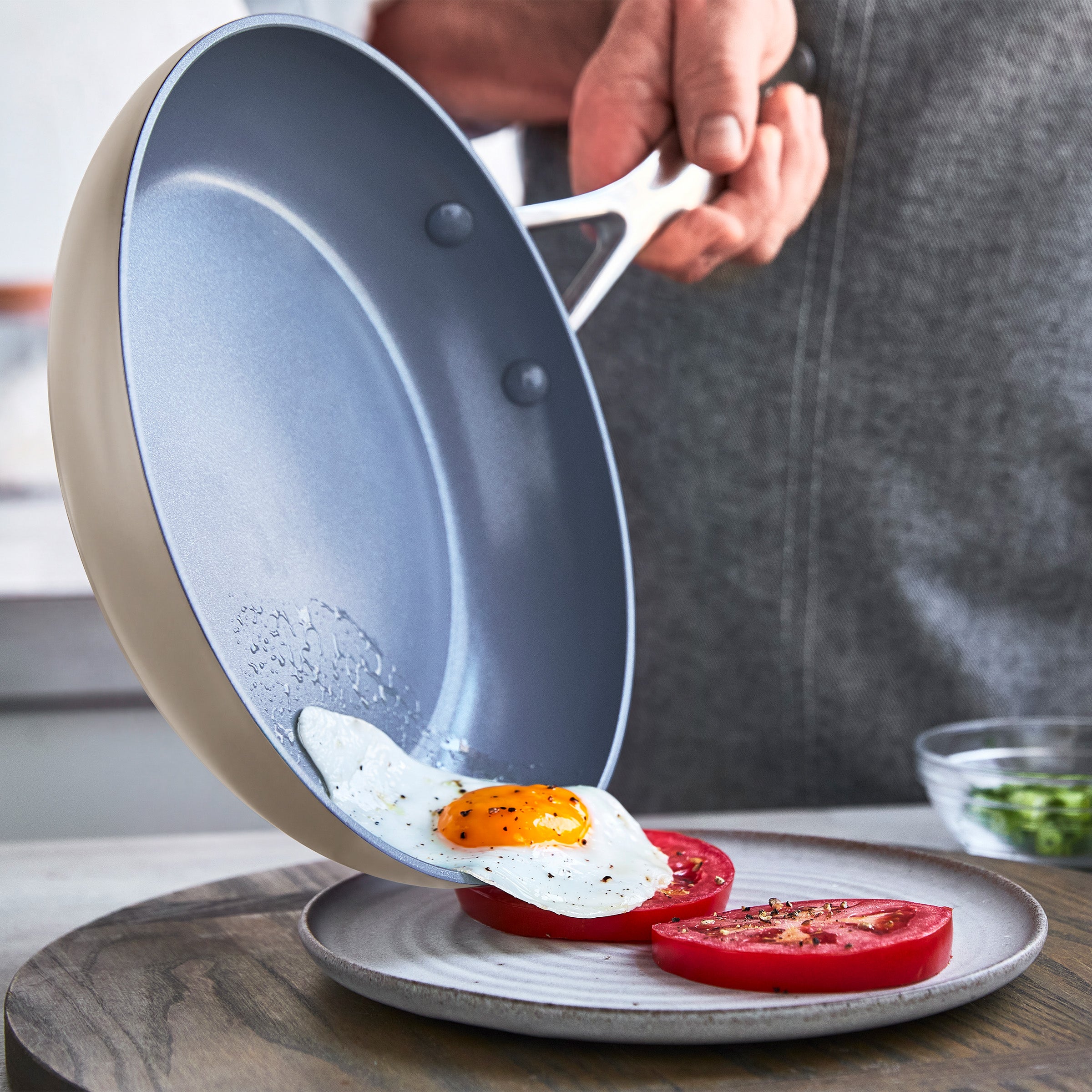 A fried egg slides from a gray ceramic nonstick frypan onto a white plate with sliced tomatoes.