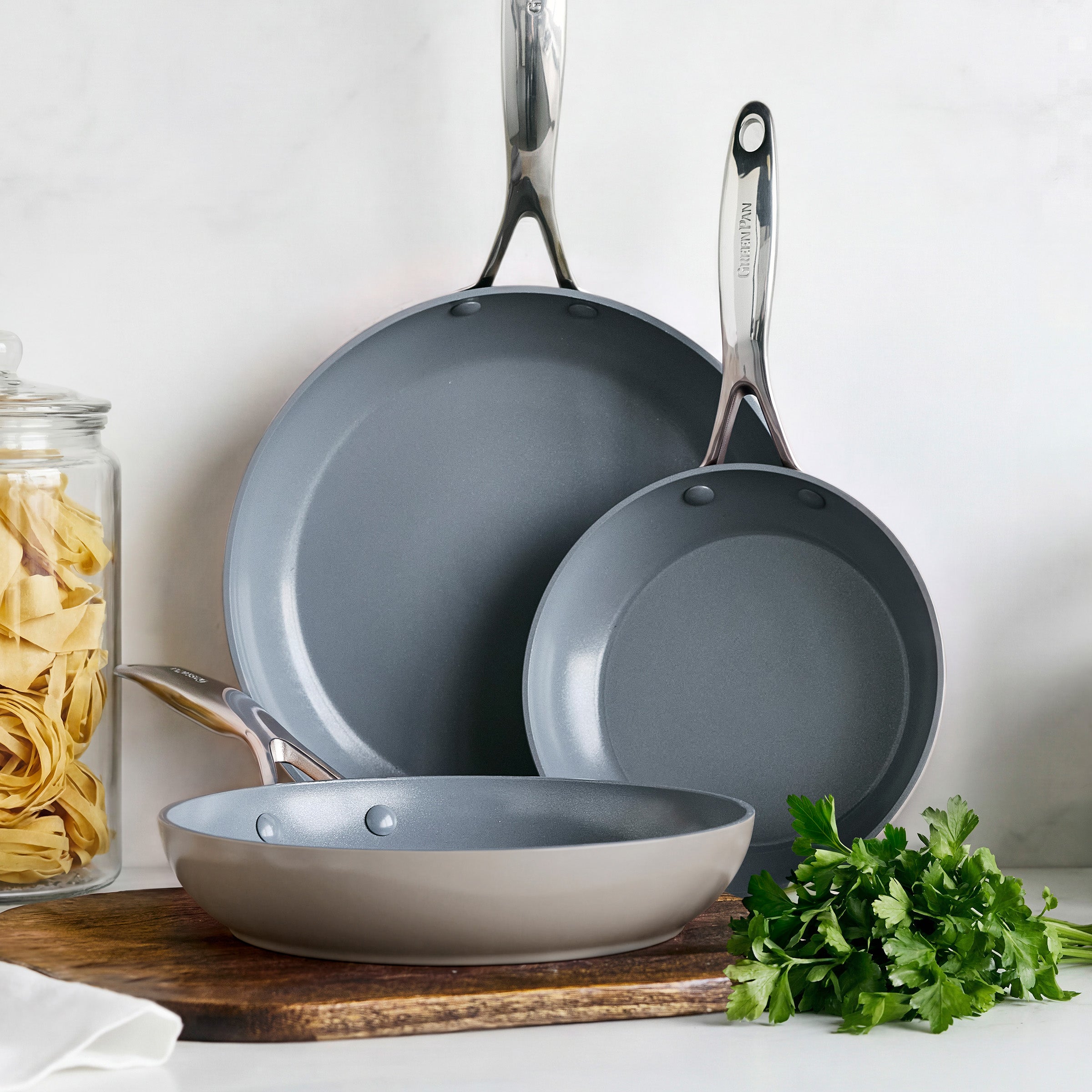 Set of three taupe ceramic nonstick frypans with metal handles on a board, parsley and a jar of pasta nearby.
