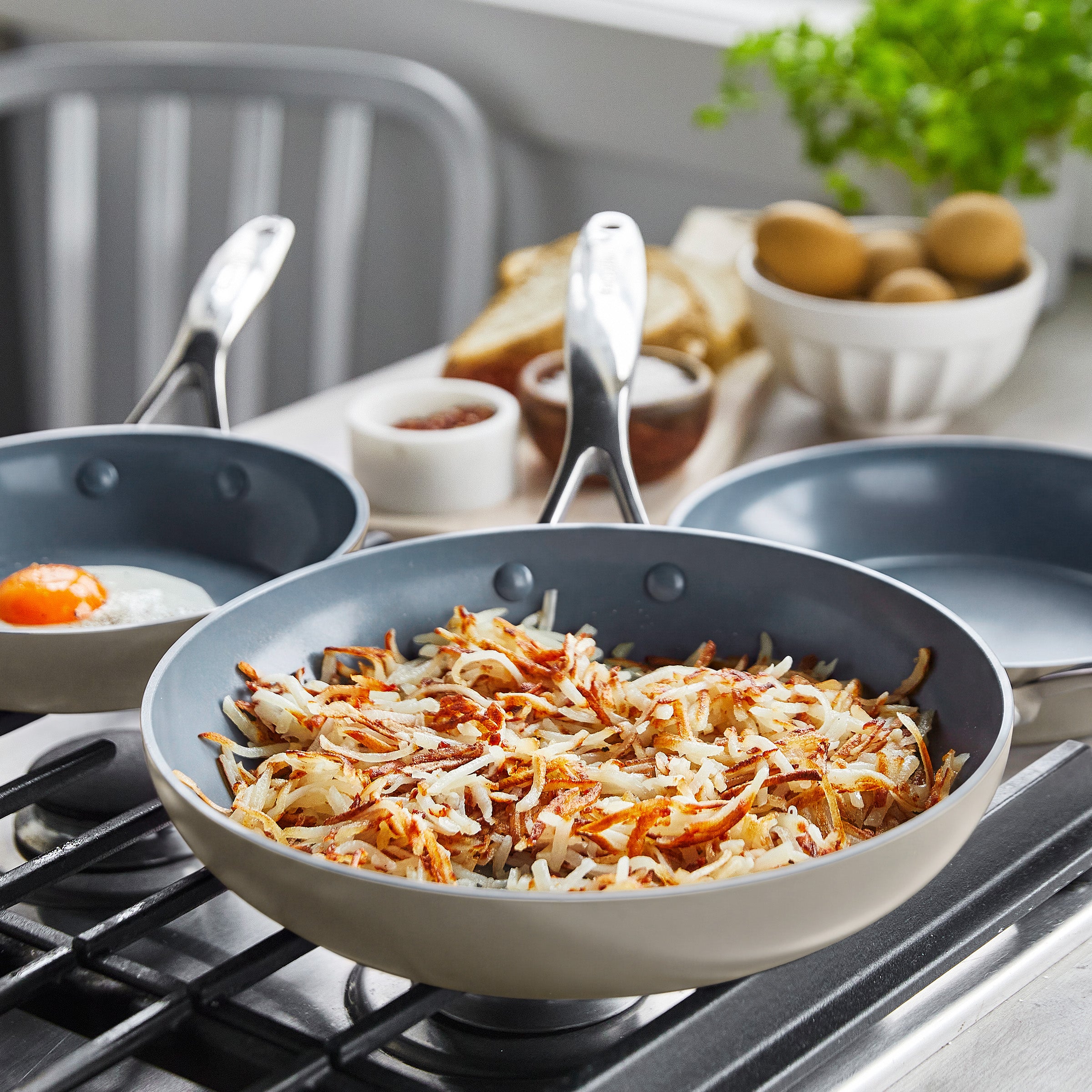 Crispy hash browns and eggs cooking in a ceramic nonstick frypan set, with fresh ingredients in the background.