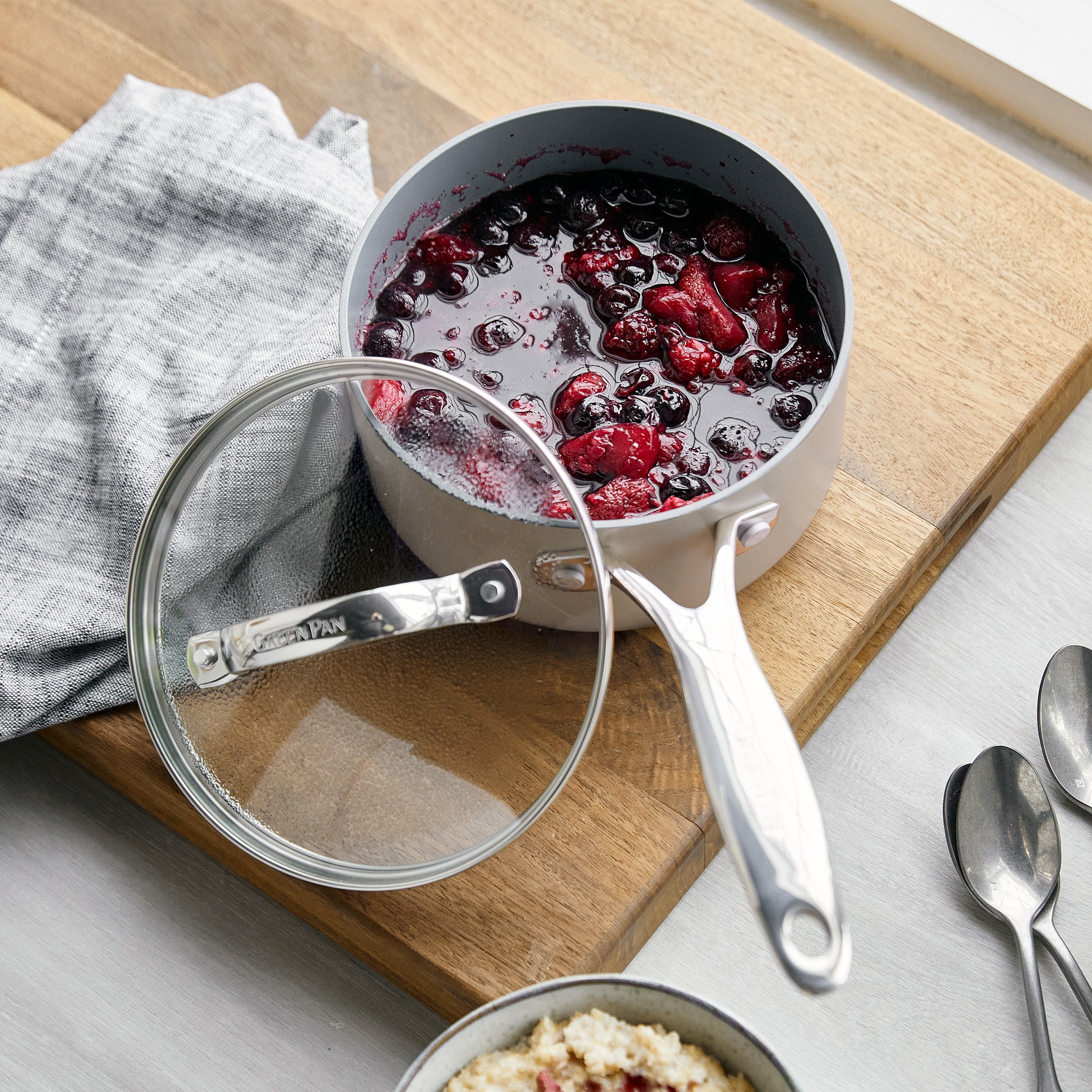 Ceramic nonstick saucepan set on a wooden board with berry compote, matching lid, gray cloth, and spoons.