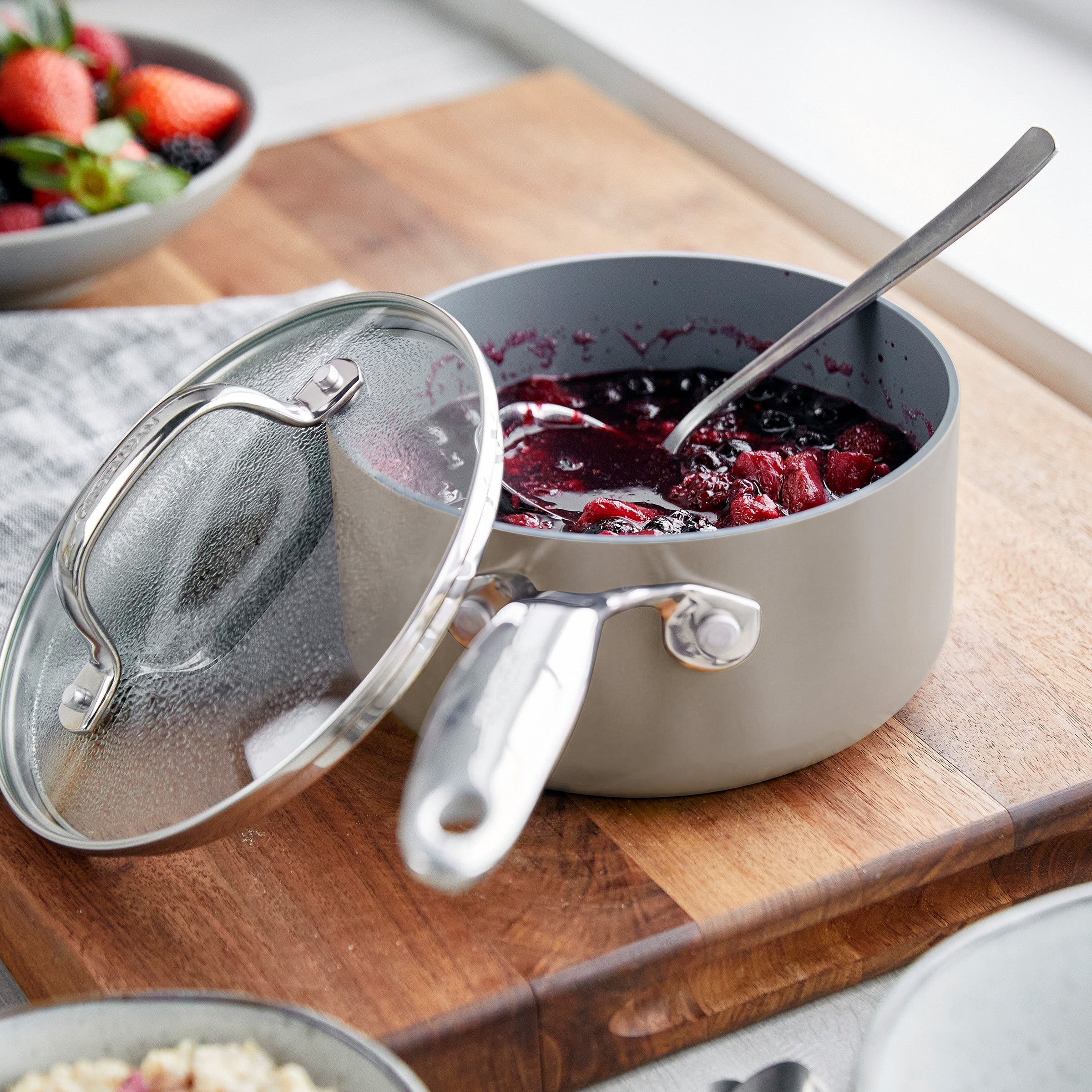 Ceramic saucepan set with berry compote inside, on a board with a spoon; matching lid is placed next to the saucepans.