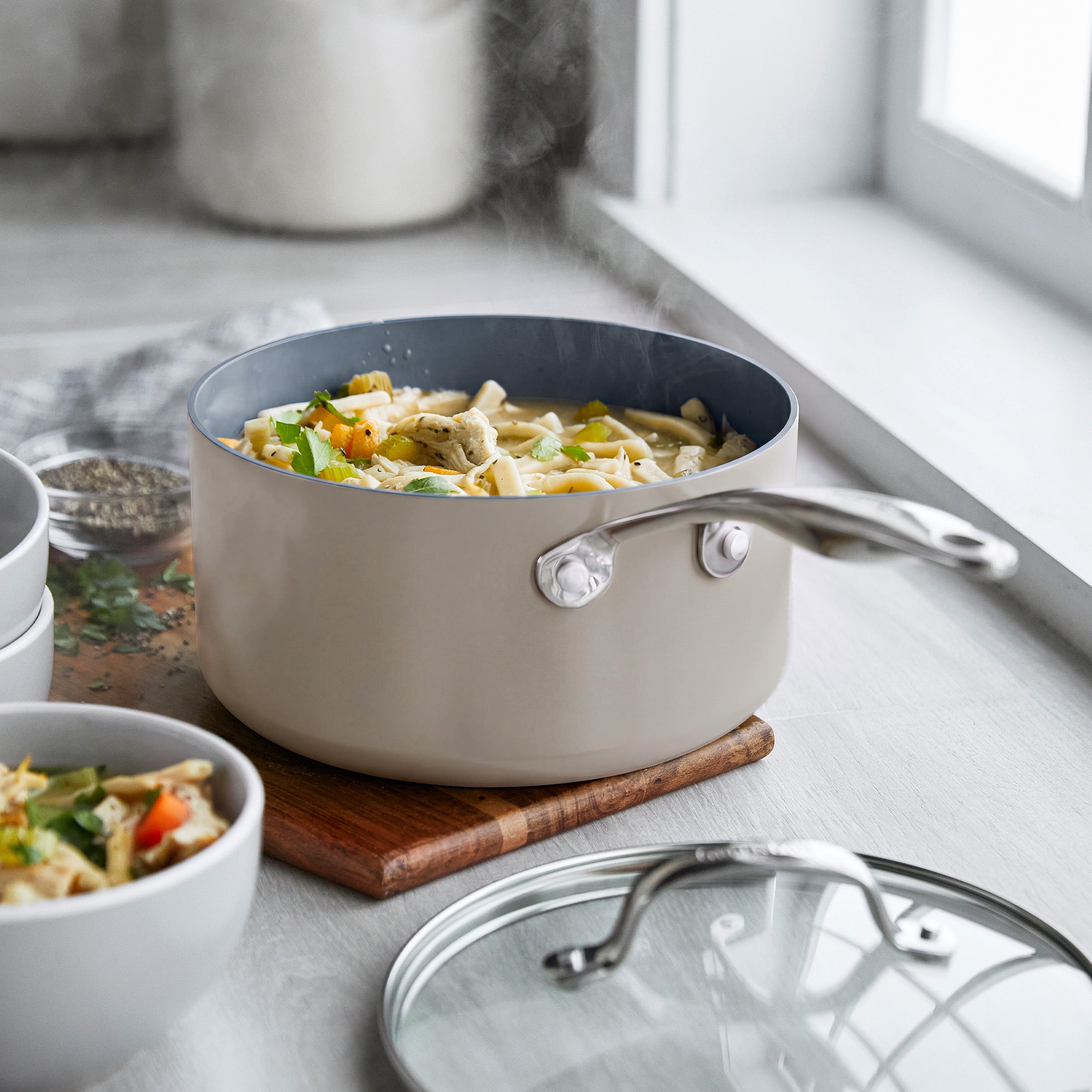 Ceramic nonstick saucepan set with lids, filled with pasta and veggies, on a wooden board by a window.