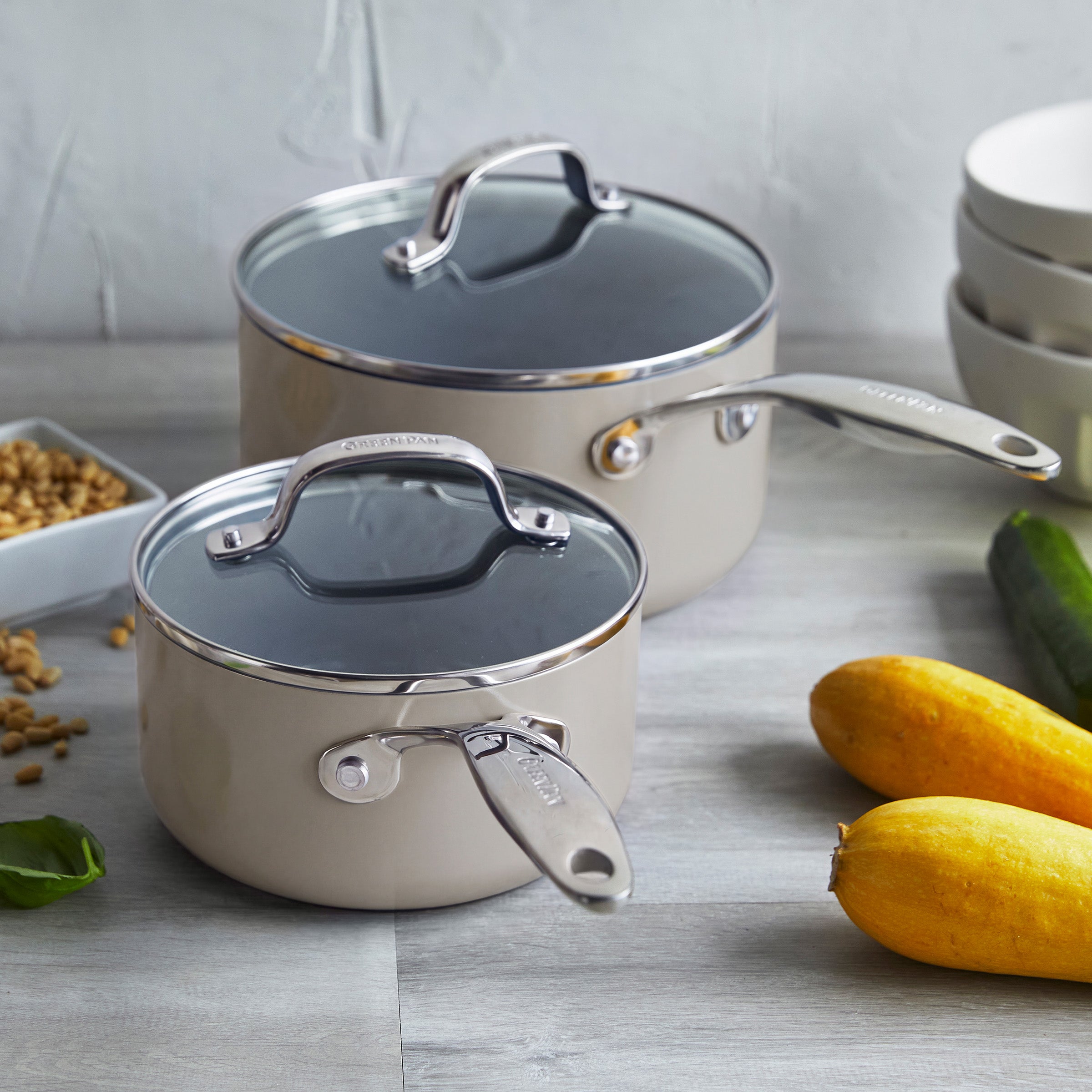 Two taupe ceramic nonstick saucepans with lids on a gray countertop, next to squash, cucumber, and bowls.