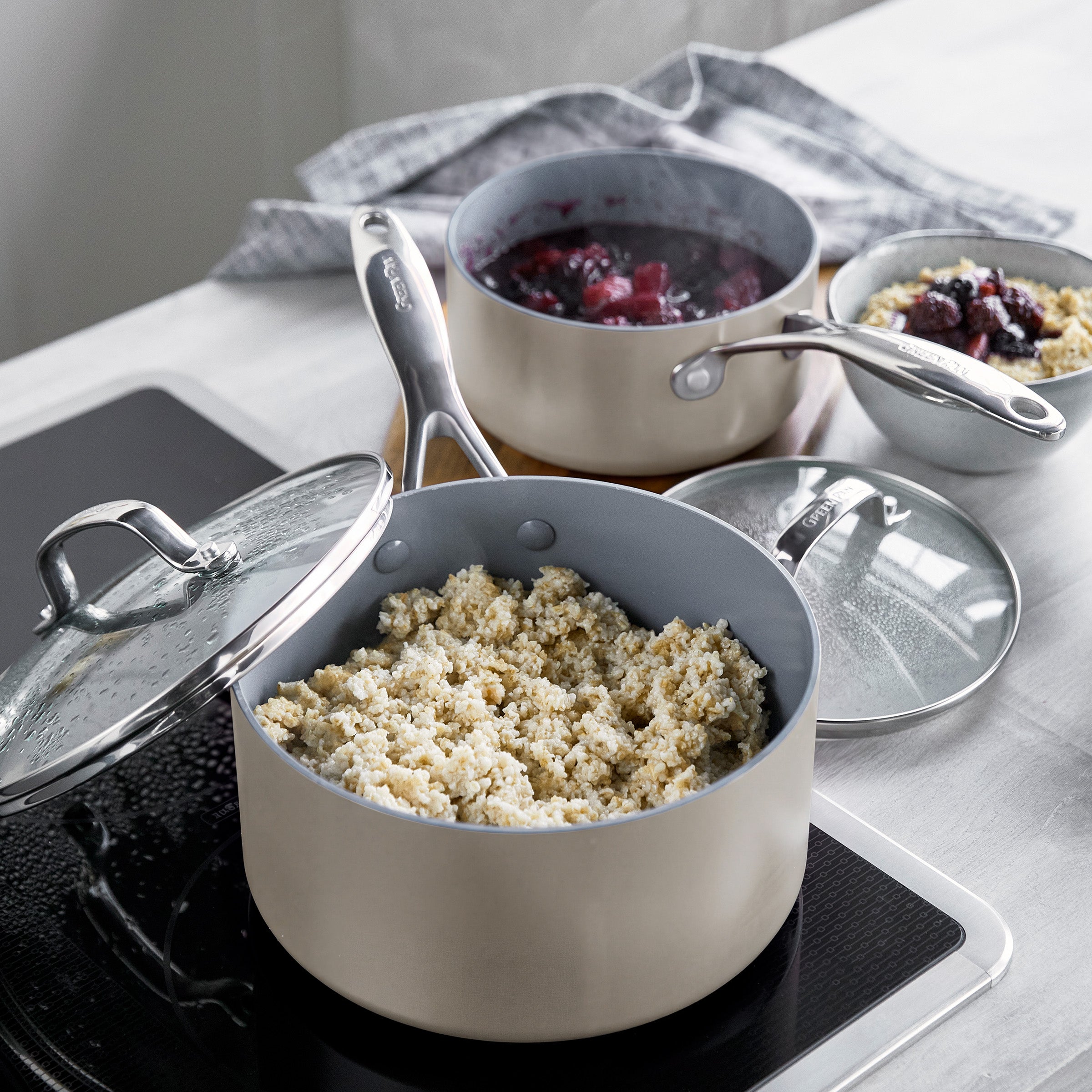 Set of two grey ceramic nonstick saucepans with lids on stove, with a grains dish and fruit bowl beside them.