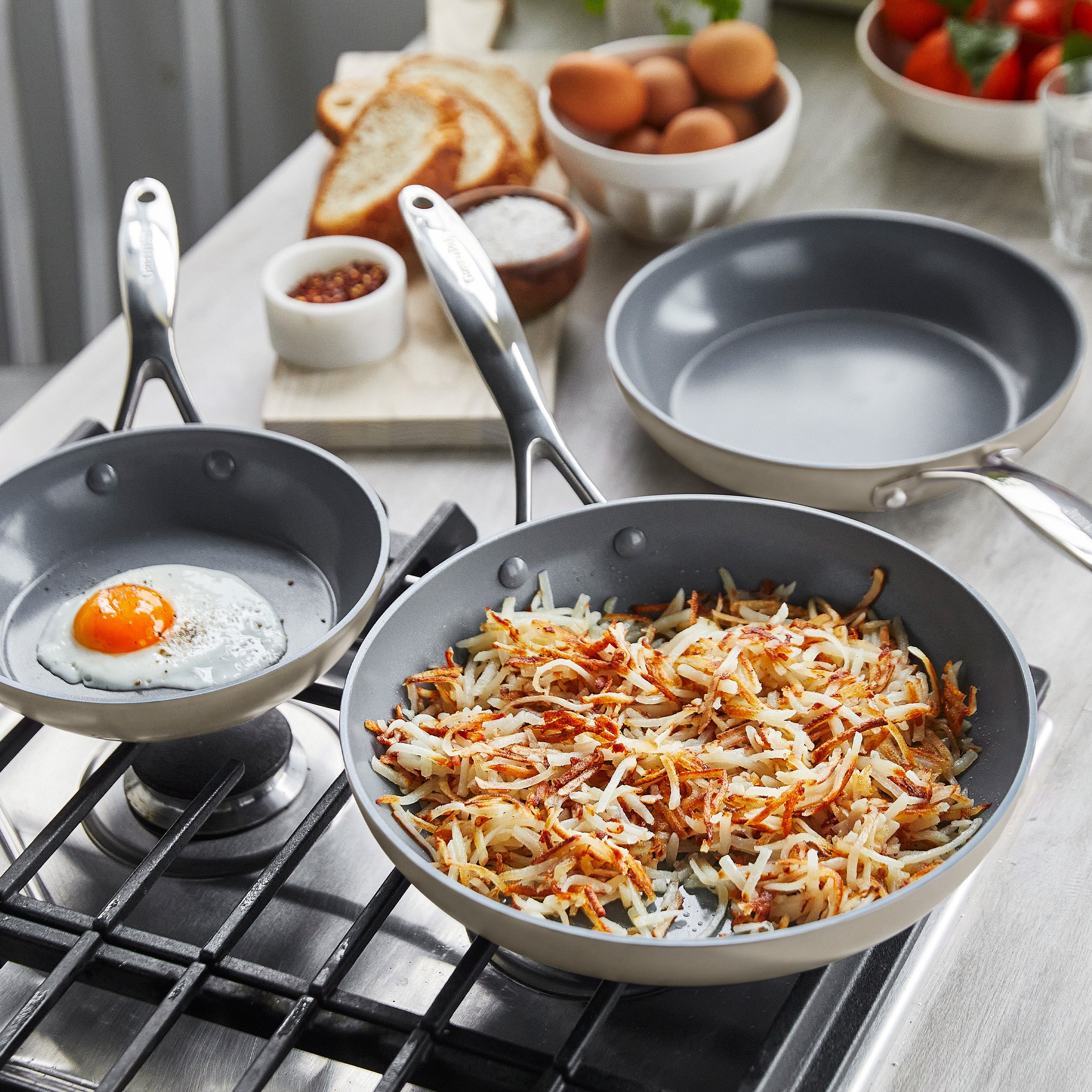 Fried egg and hash browns in gray ceramic nonstick pans on the stove, with bread, eggs, and tomatoes on the counter nearby.