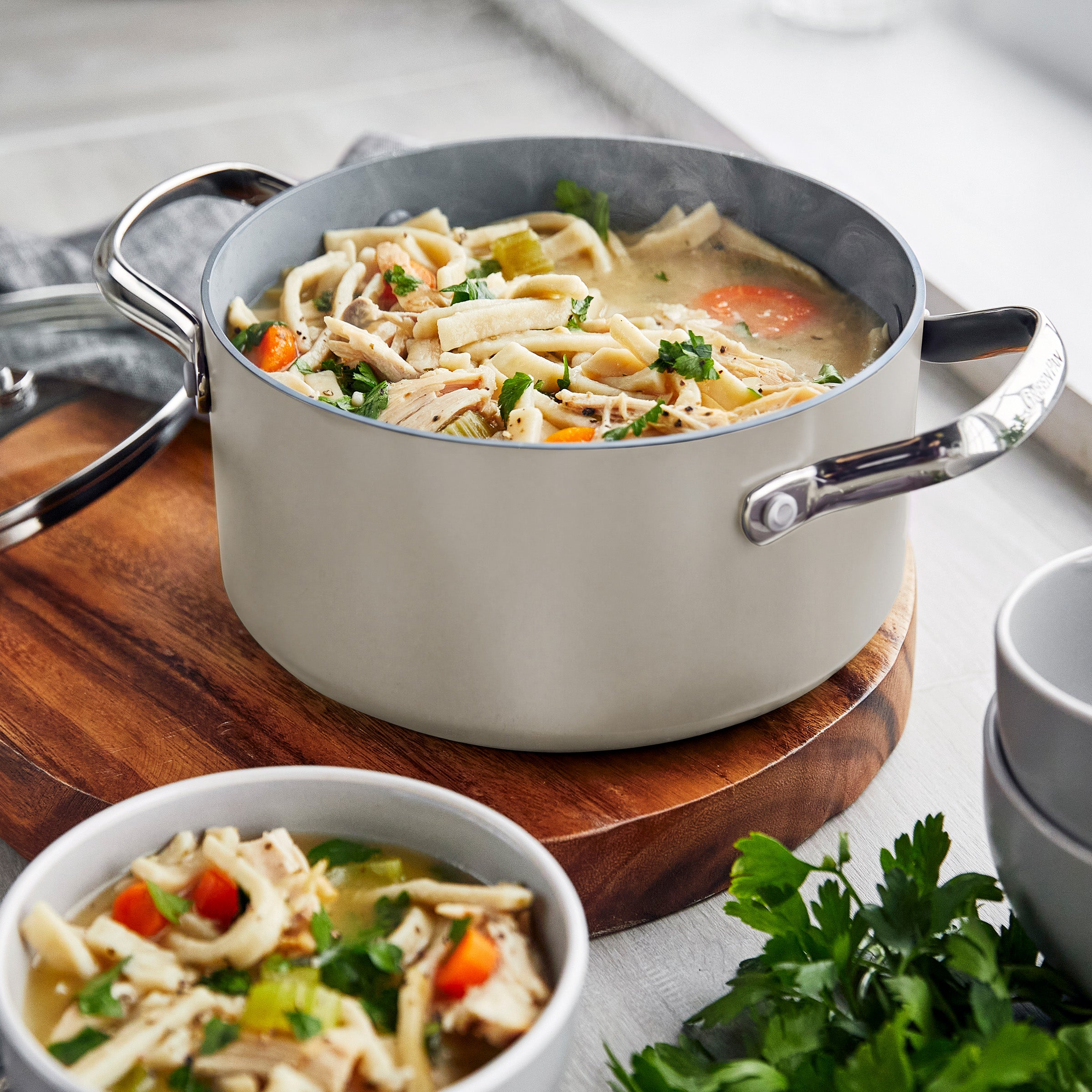 Veggie noodle soup in a white ceramic nonstick pot on the stove, with a nearby bowl and fresh parsley.