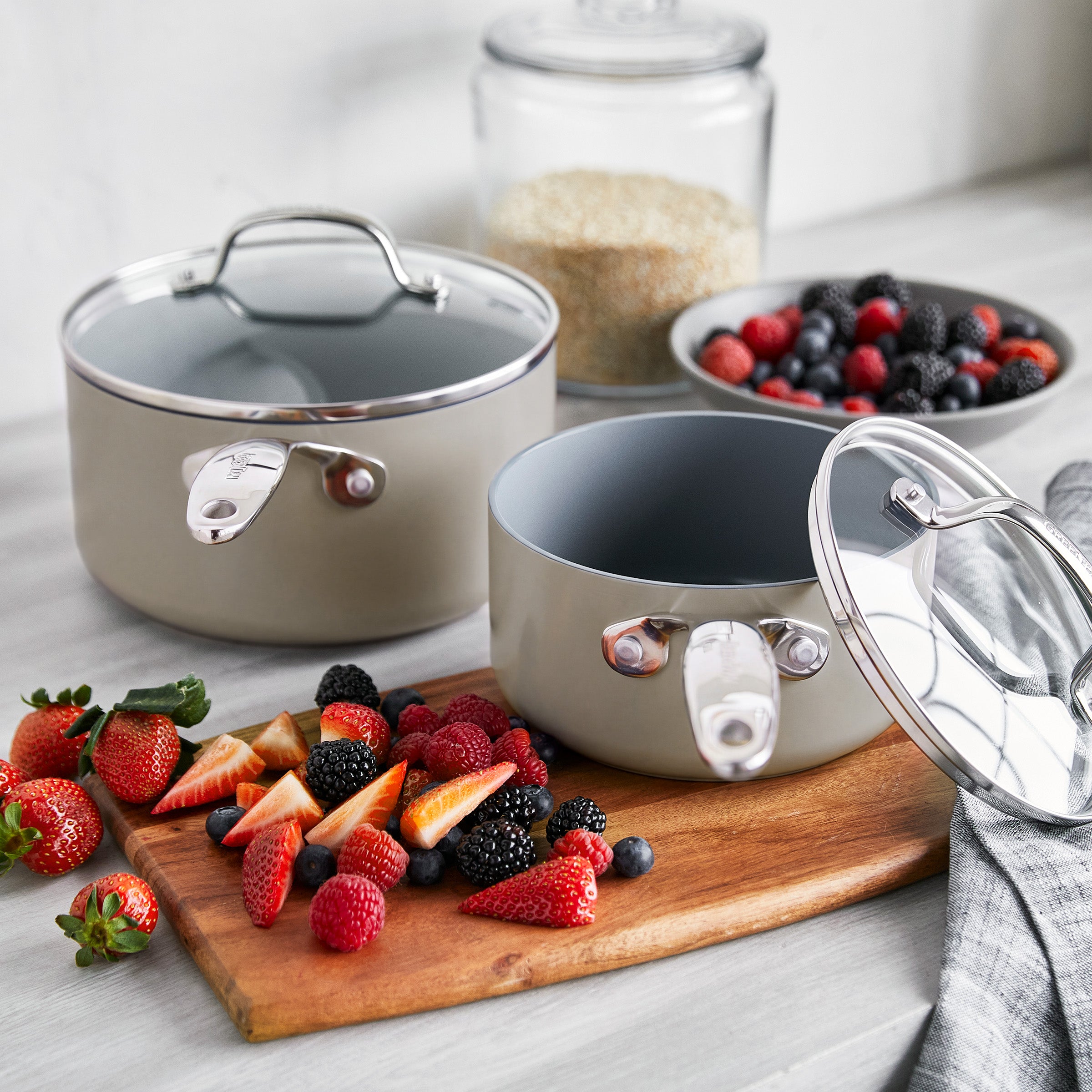 Taupe ceramic nonstick cookware set with glass lids on a kitchen counter beside bowls of berries and grains.