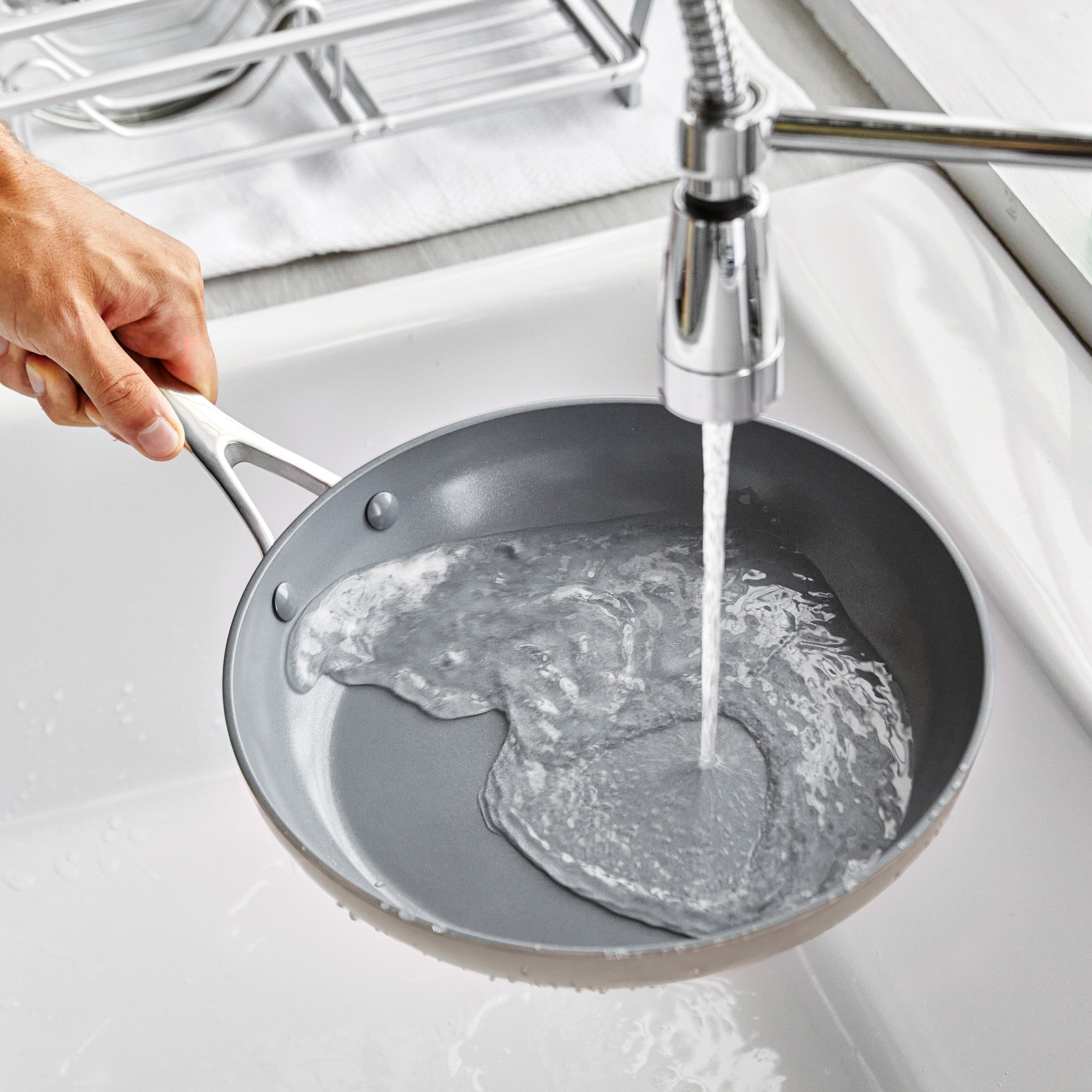 Hand rinsing a taupe ceramic nonstick pan from a 19-piece set under running water in a kitchen sink.
