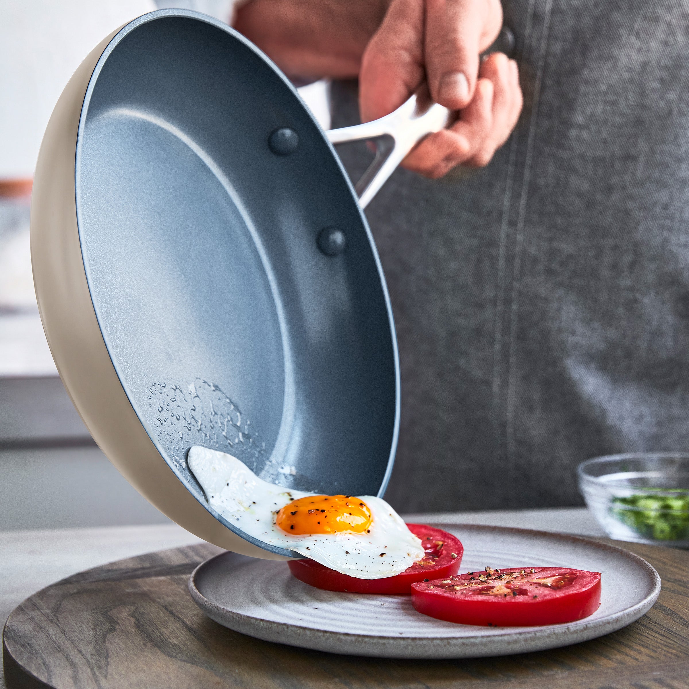 Fried egg being slid from a ceramic nonstick pan onto tomato slices on a white plate.