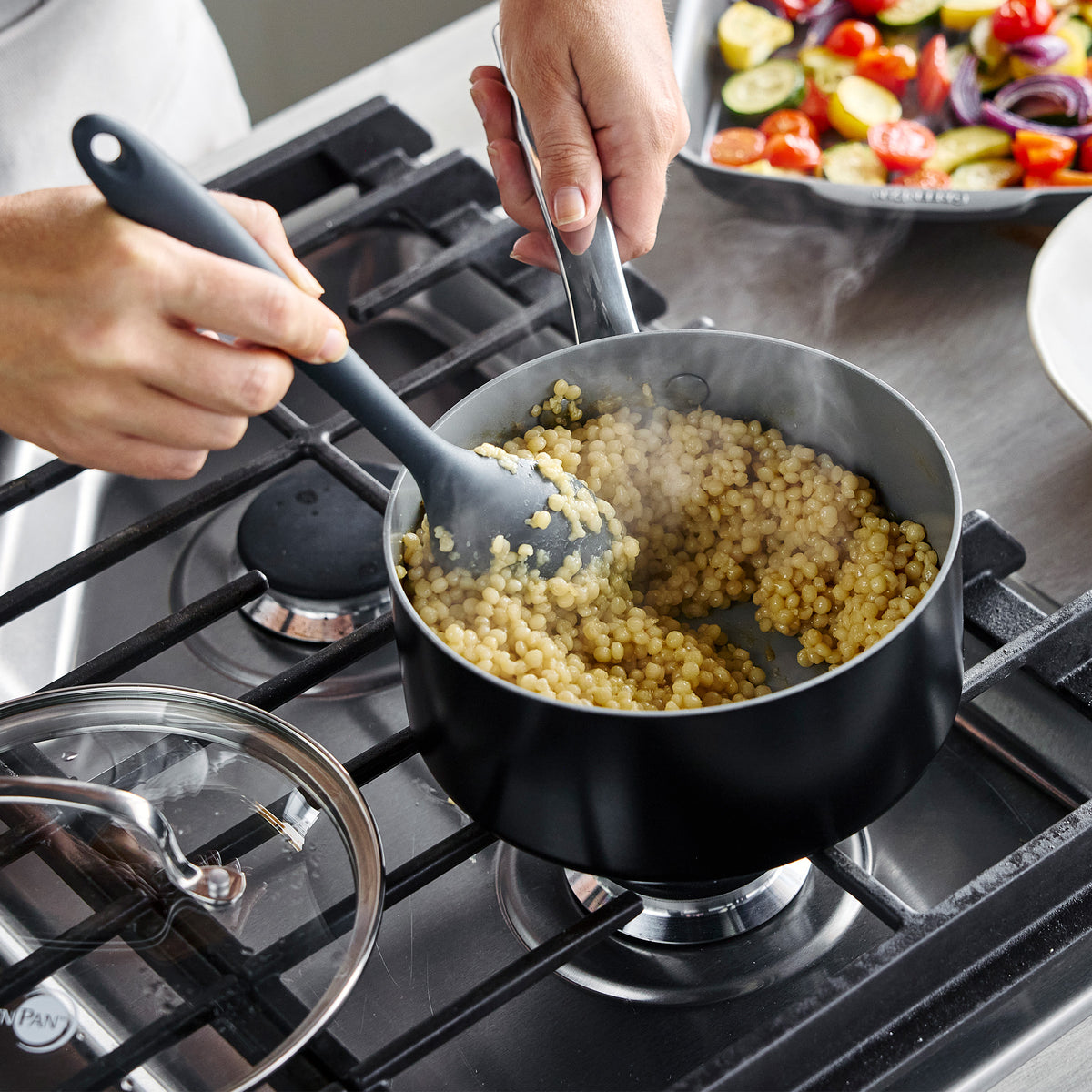 Steaming couscous being stirred in a black Chatham Pro Ceramic Nonstick 12-piece saucepan on a gas stovetop.
