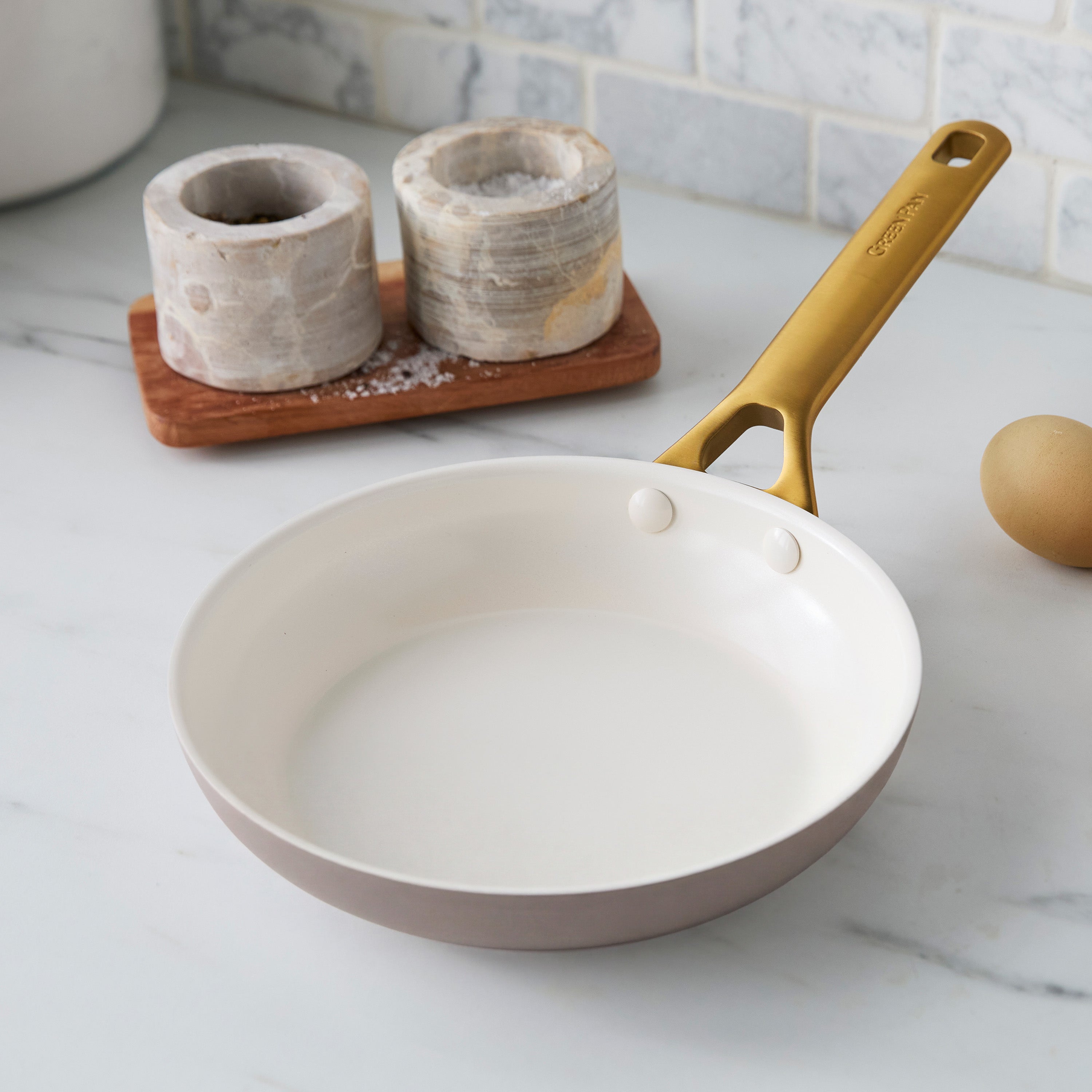 Latte ceramic nonstick 8″ frypan with yellow handle on marble, beside two salt cellars and a brown egg.