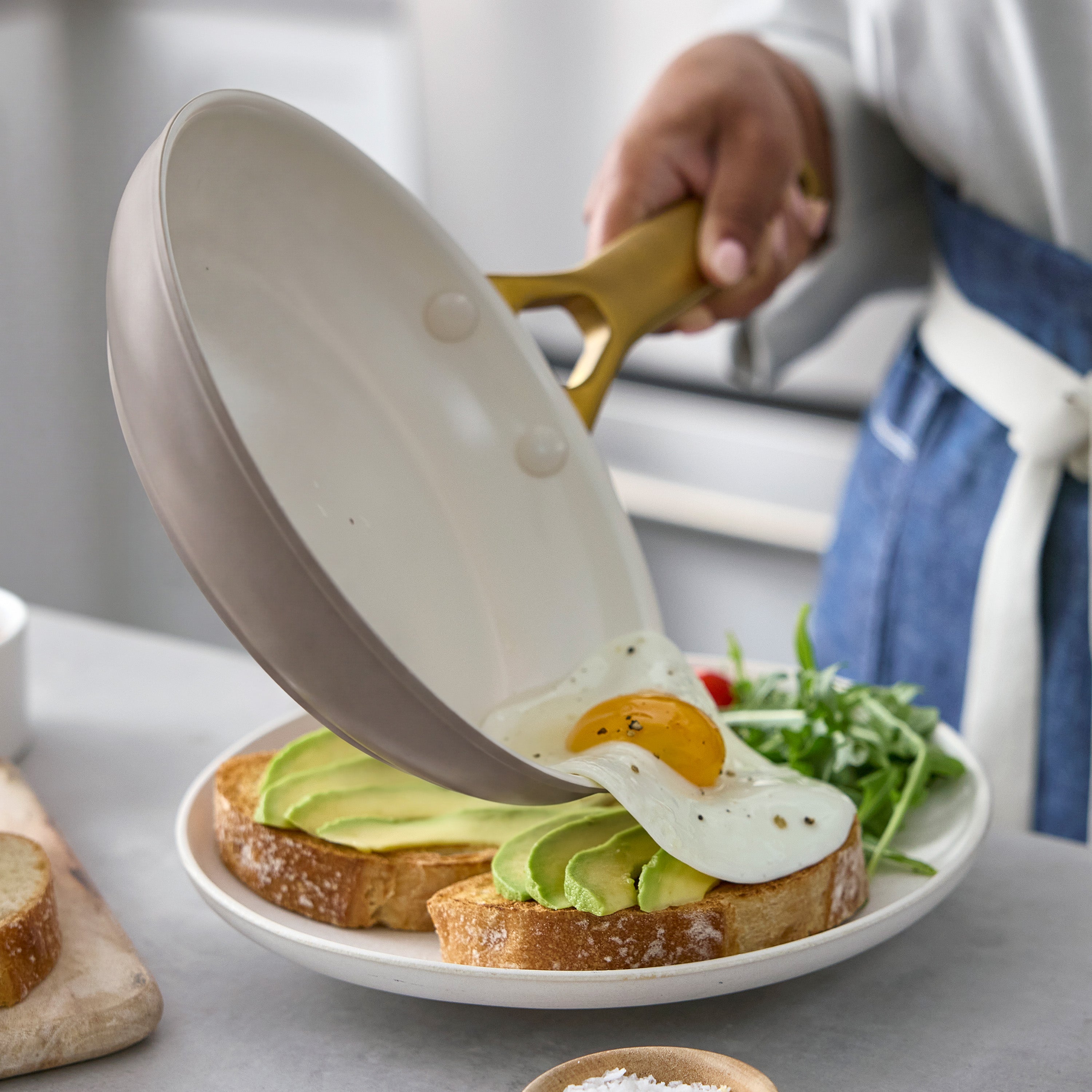 A fried egg being slid from a light-colored ceramic nonstick frypan onto avocado toast with greens.