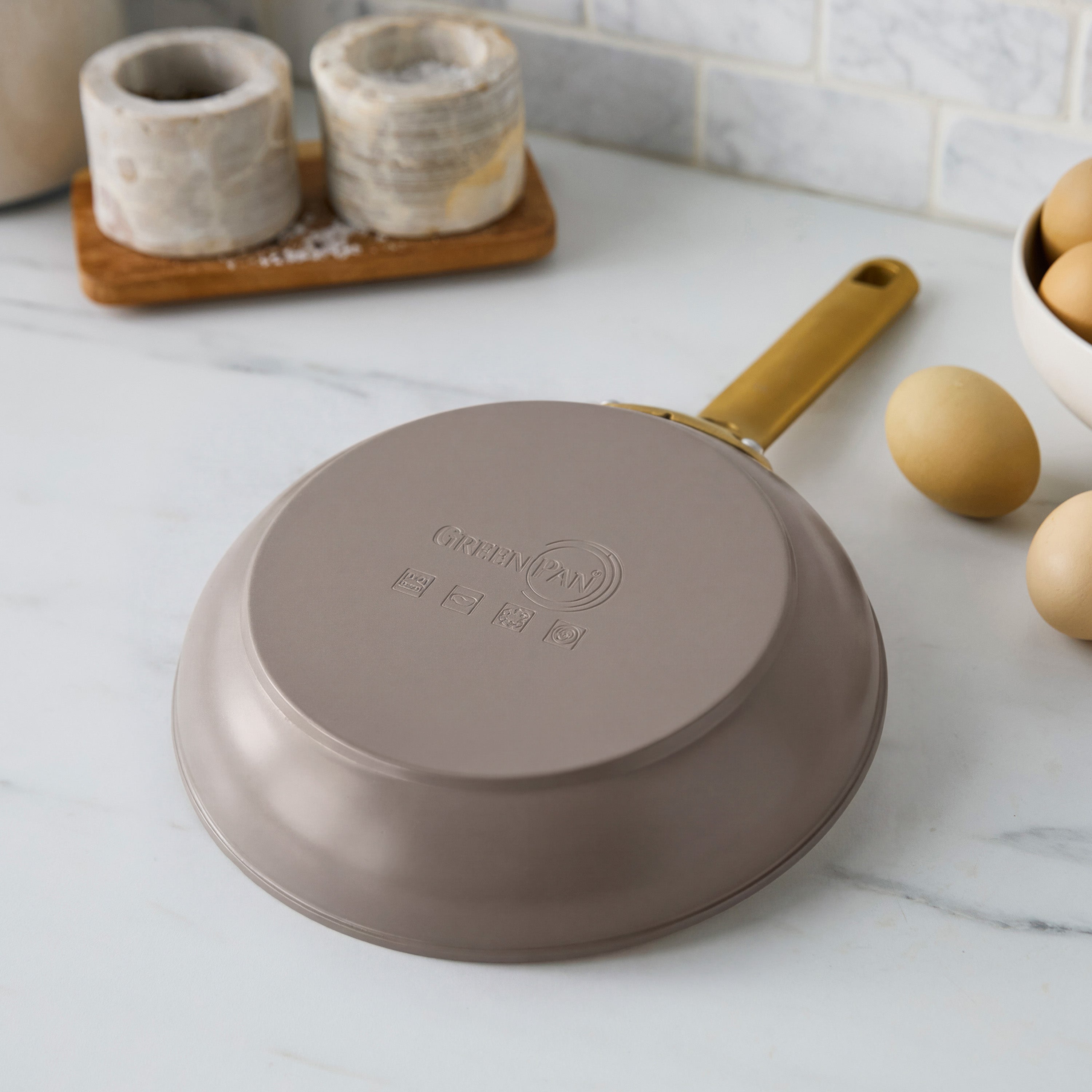 Latte-colored ceramic nonstick 8″ frypan with gold handle upside down on white counter near eggs and kitchen containers.