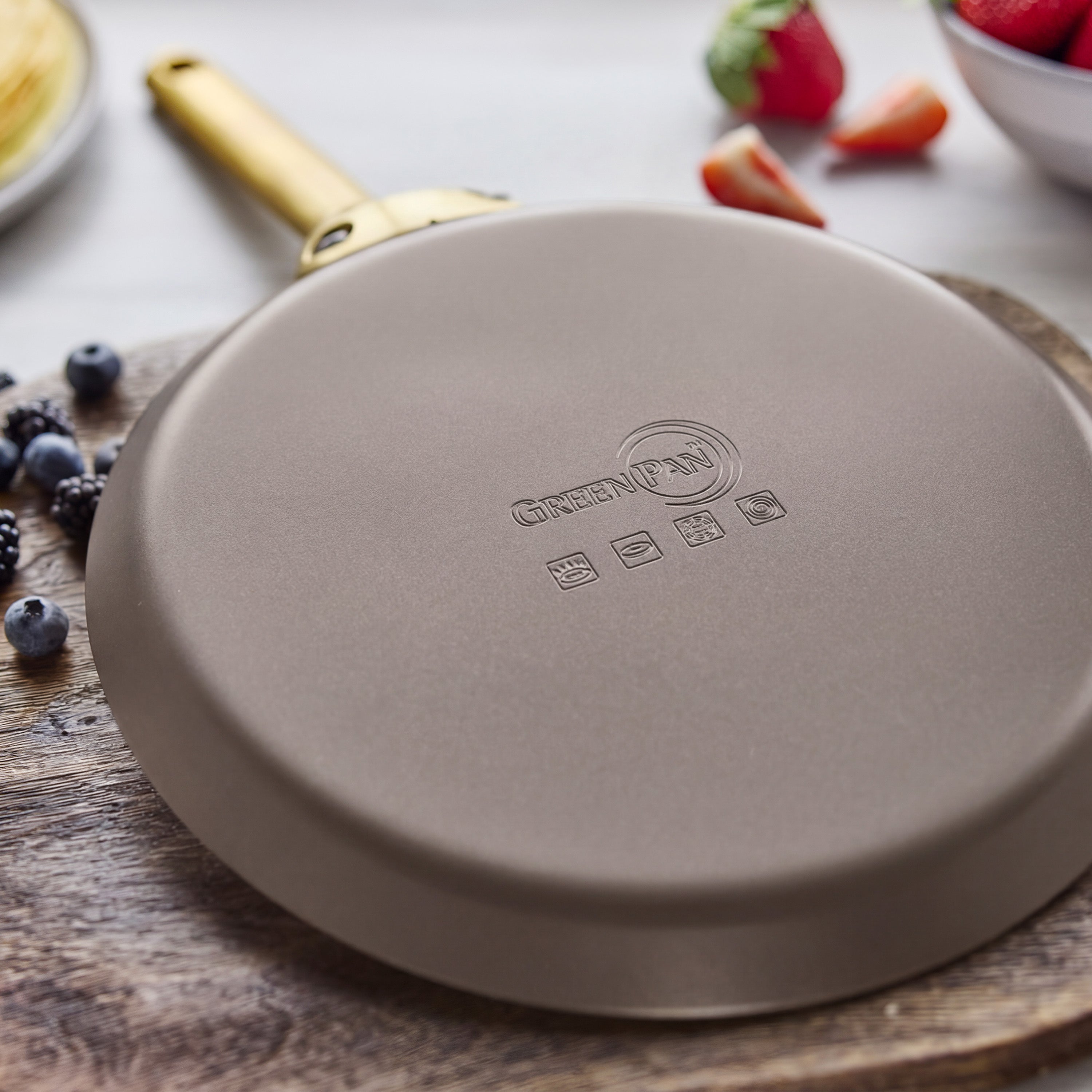 Upside down ceramic nonstick crepe pan in latte color on a board, with berries and pancakes blurred in the background.