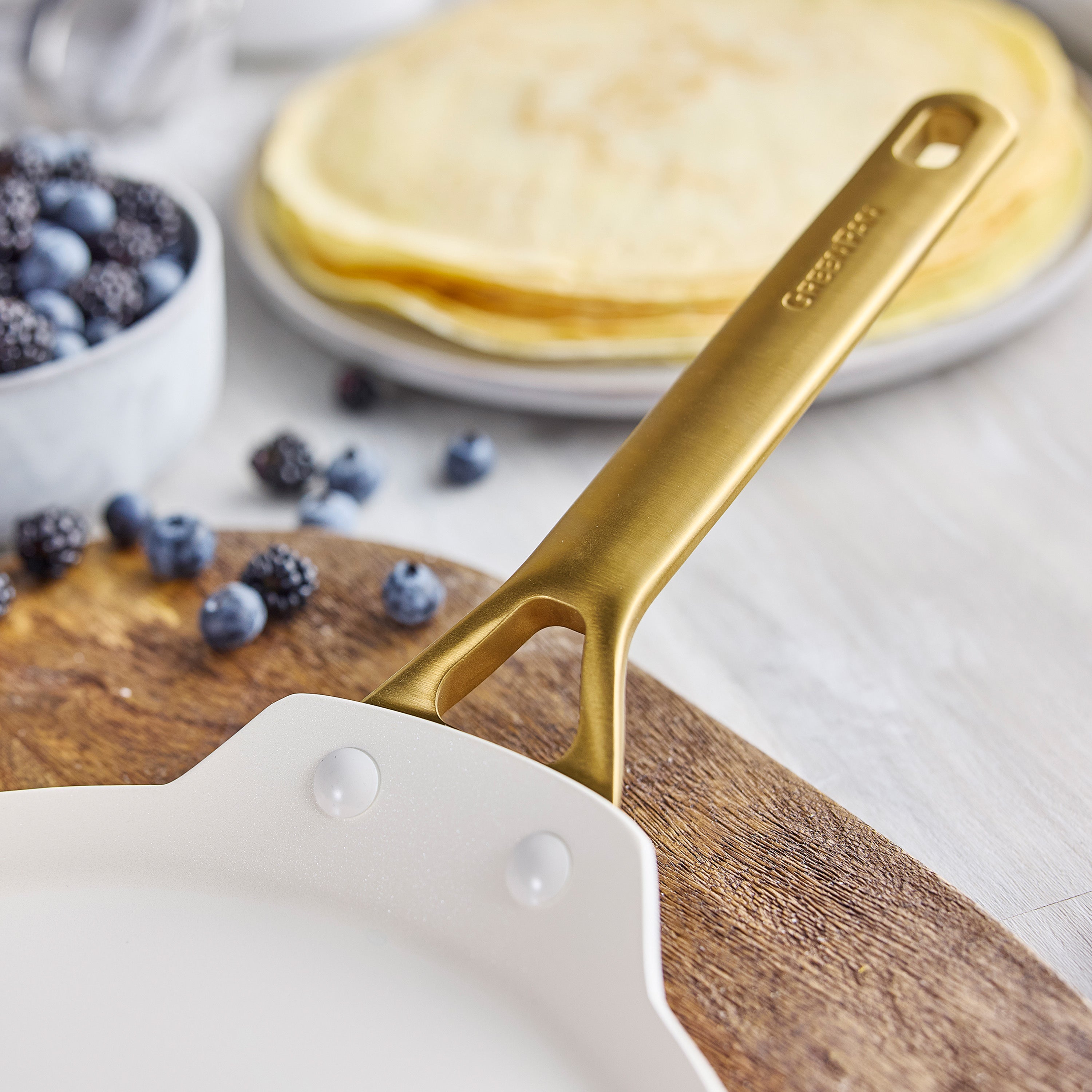 Latte ceramic crepe pan with gold handle on wood, with crepes and mixed berries in the background.