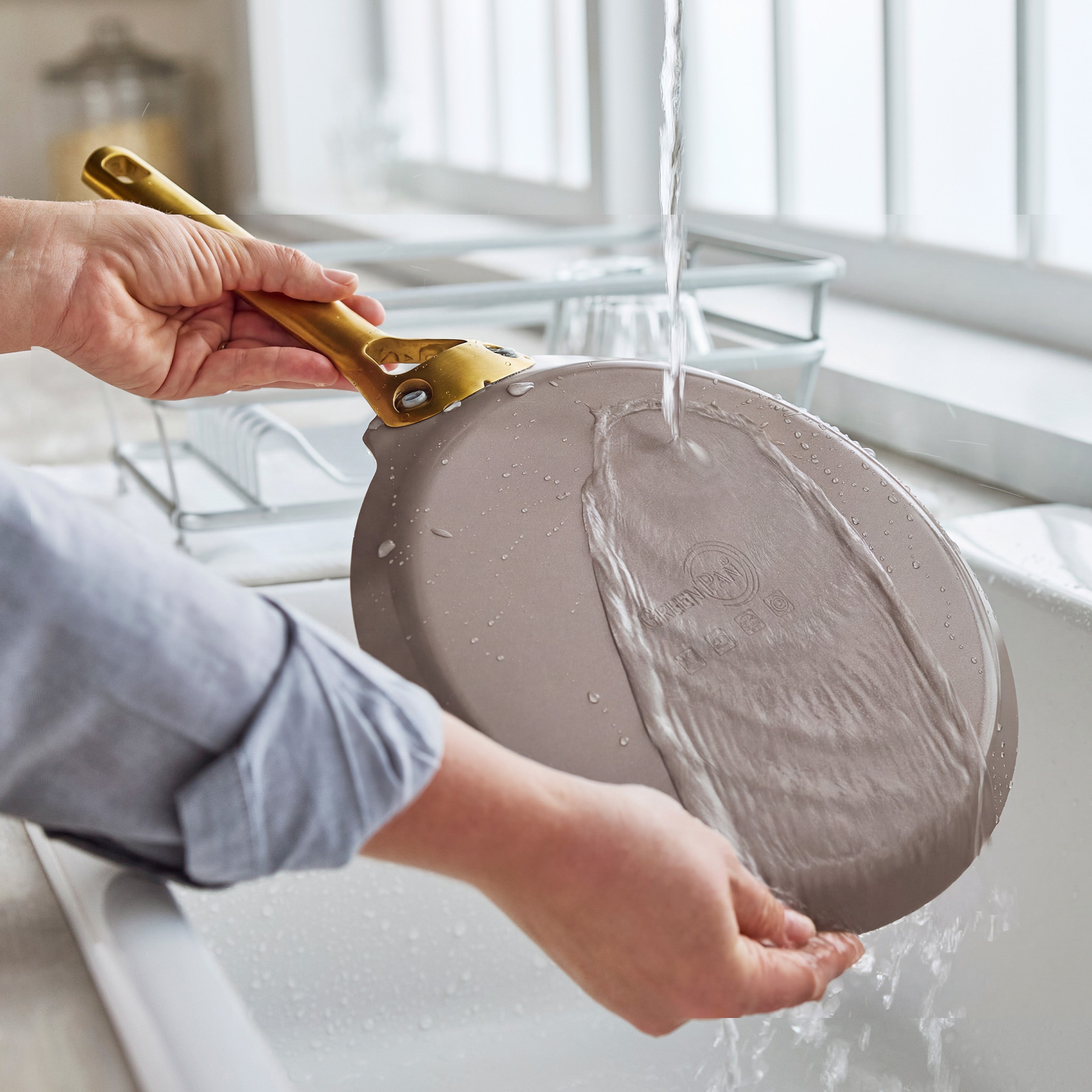 A hand rinses a cream-colored ceramic nonstick crepe pan with a gold handle under running water at a kitchen sink.