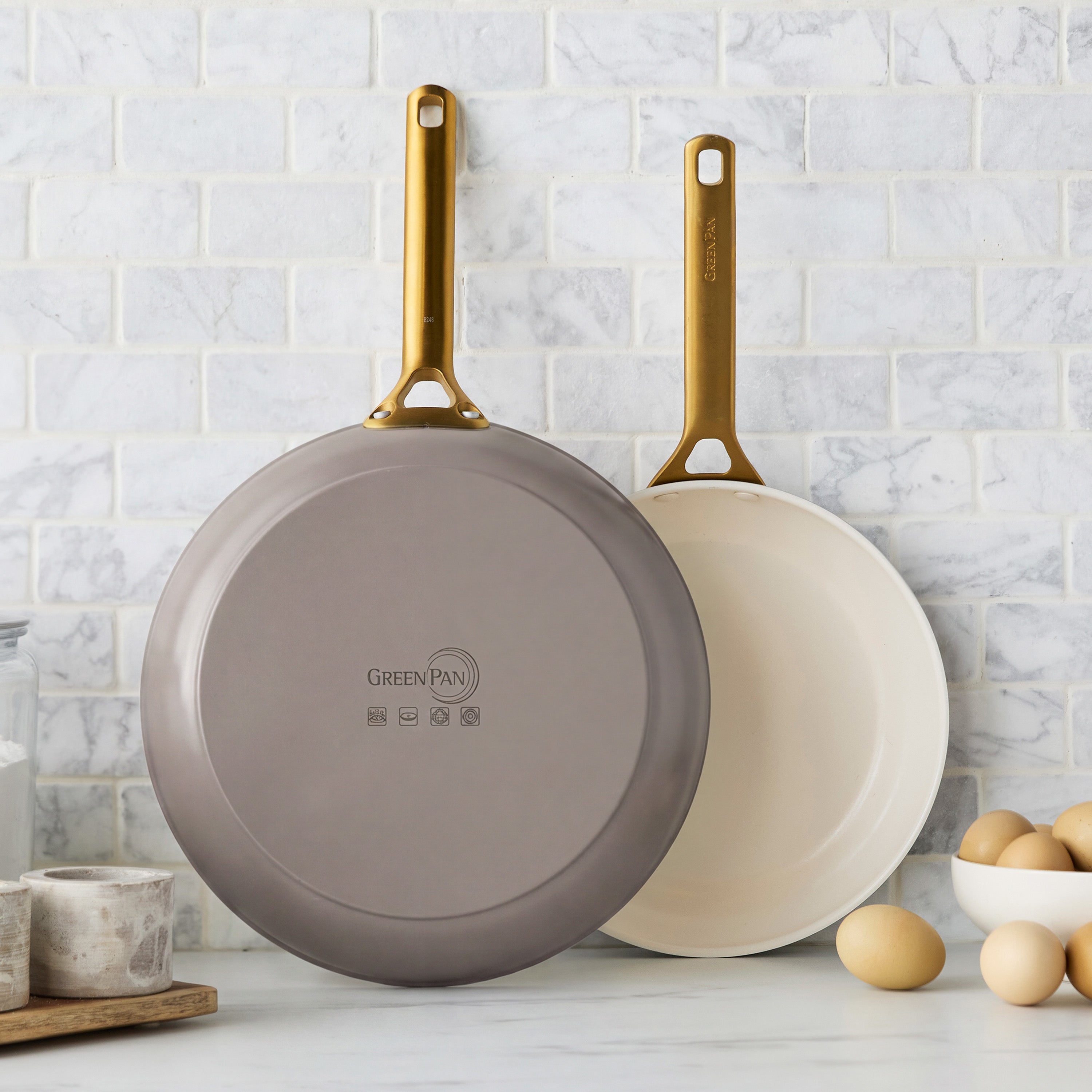 Set of two latte-colored ceramic nonstick frypans with gold handles on marble, eggs placed nearby.