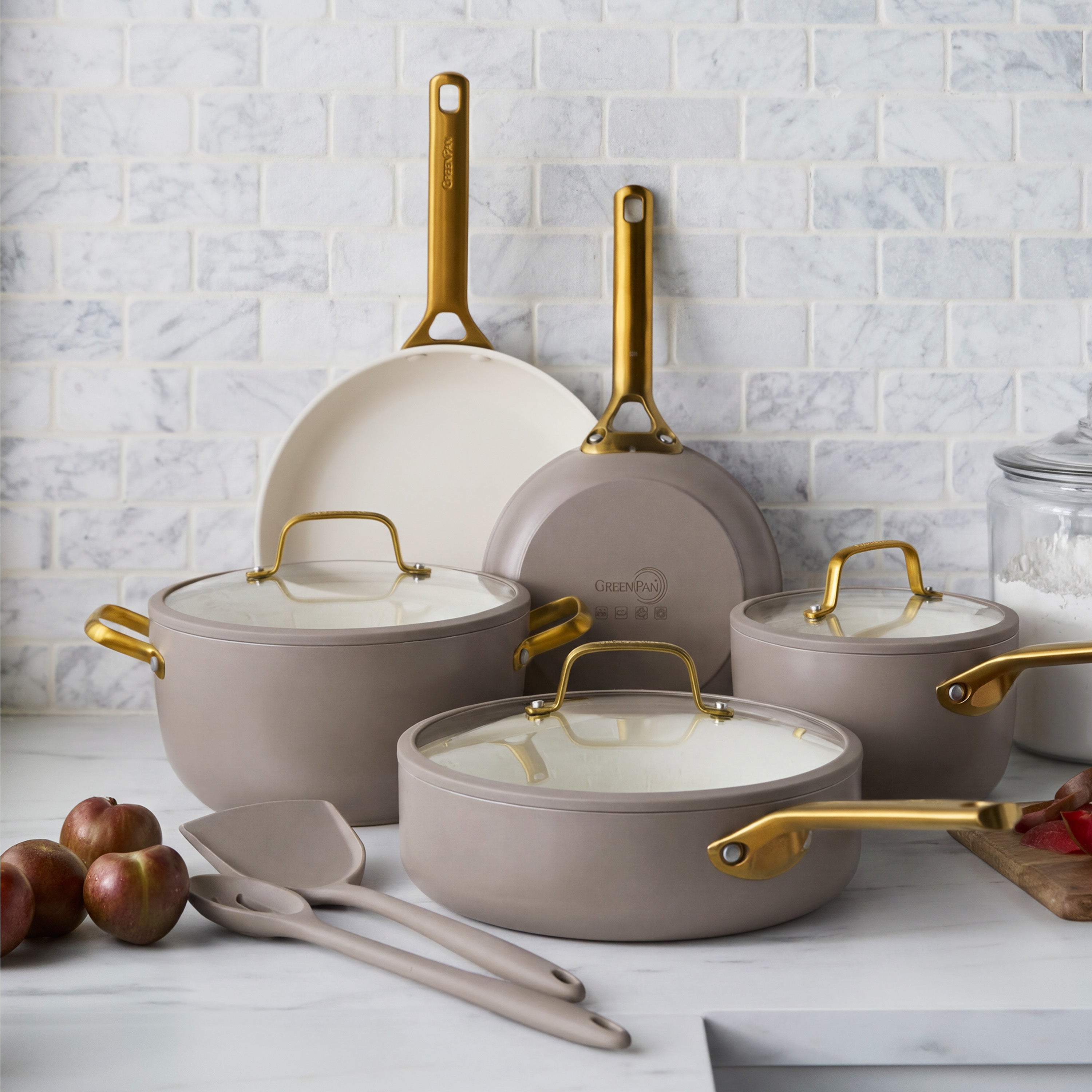 10-piece ceramic nonstick cookware set in latte with gold handles on marble, surrounded by apples and utensils.