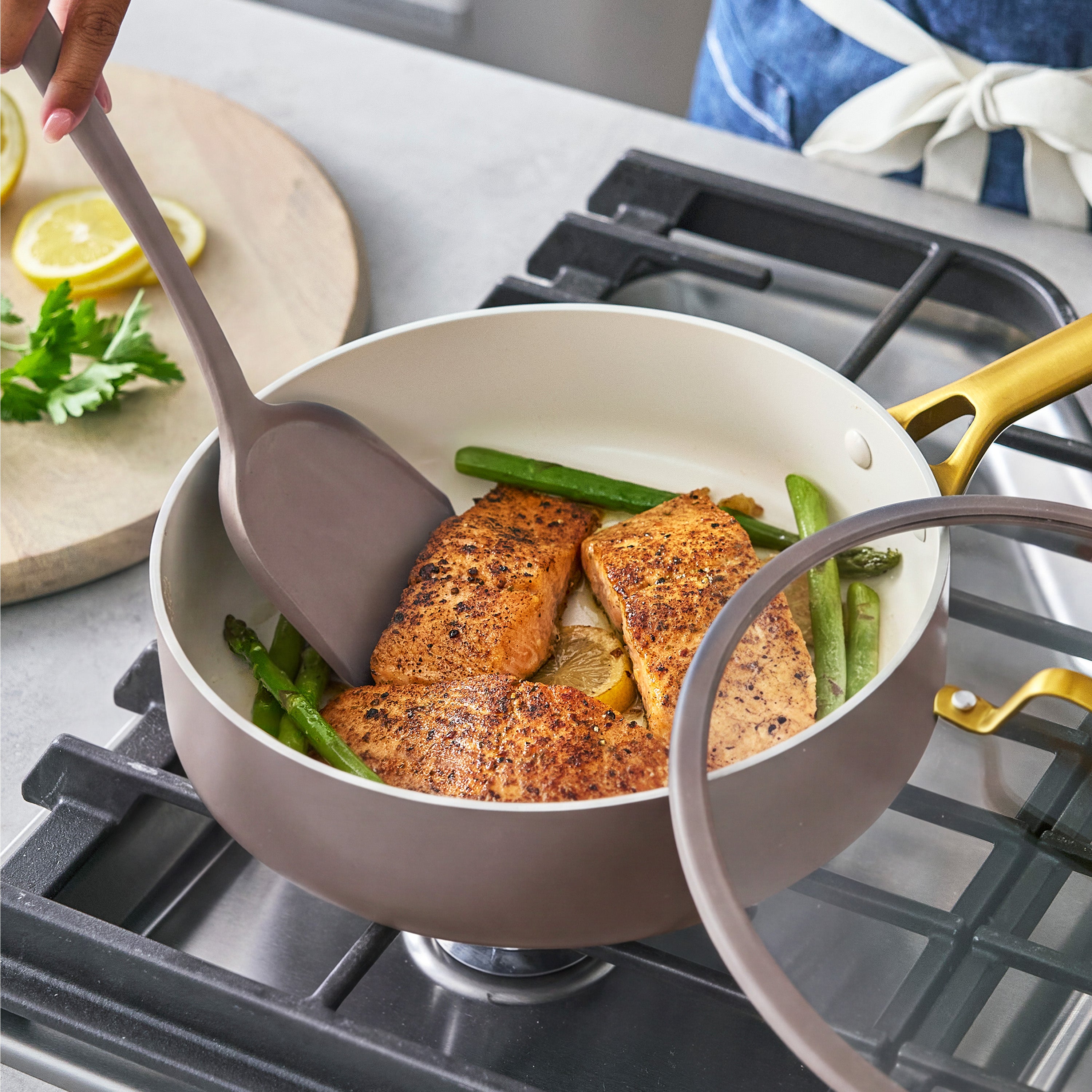 Salmon, asparagus, lemon slices, and parsley cook in a ceramic nonstick pan set as a hand stirs.