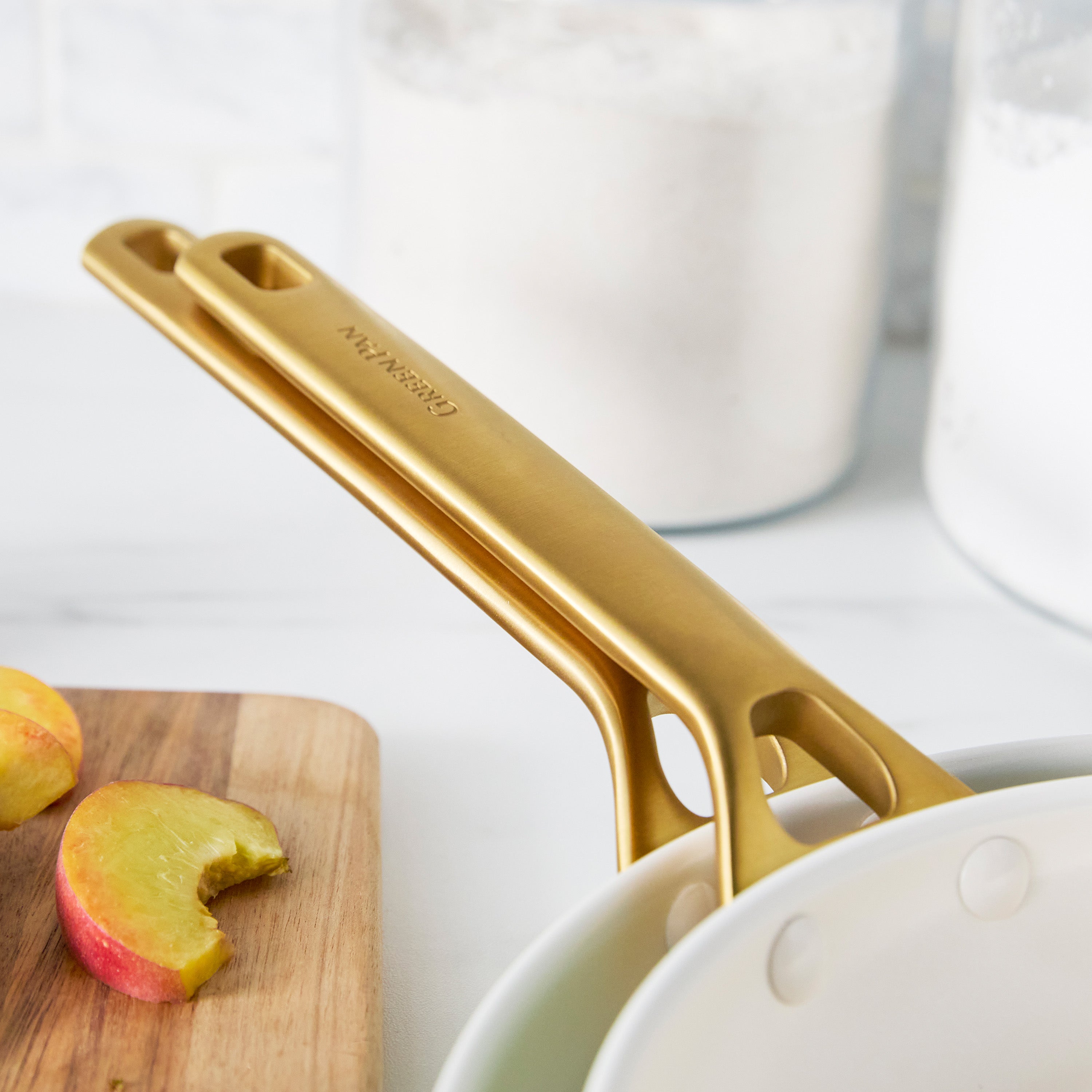 Latte-colored nonstick pan with a gold handle, peach slices inside, and glass jars in the background.
