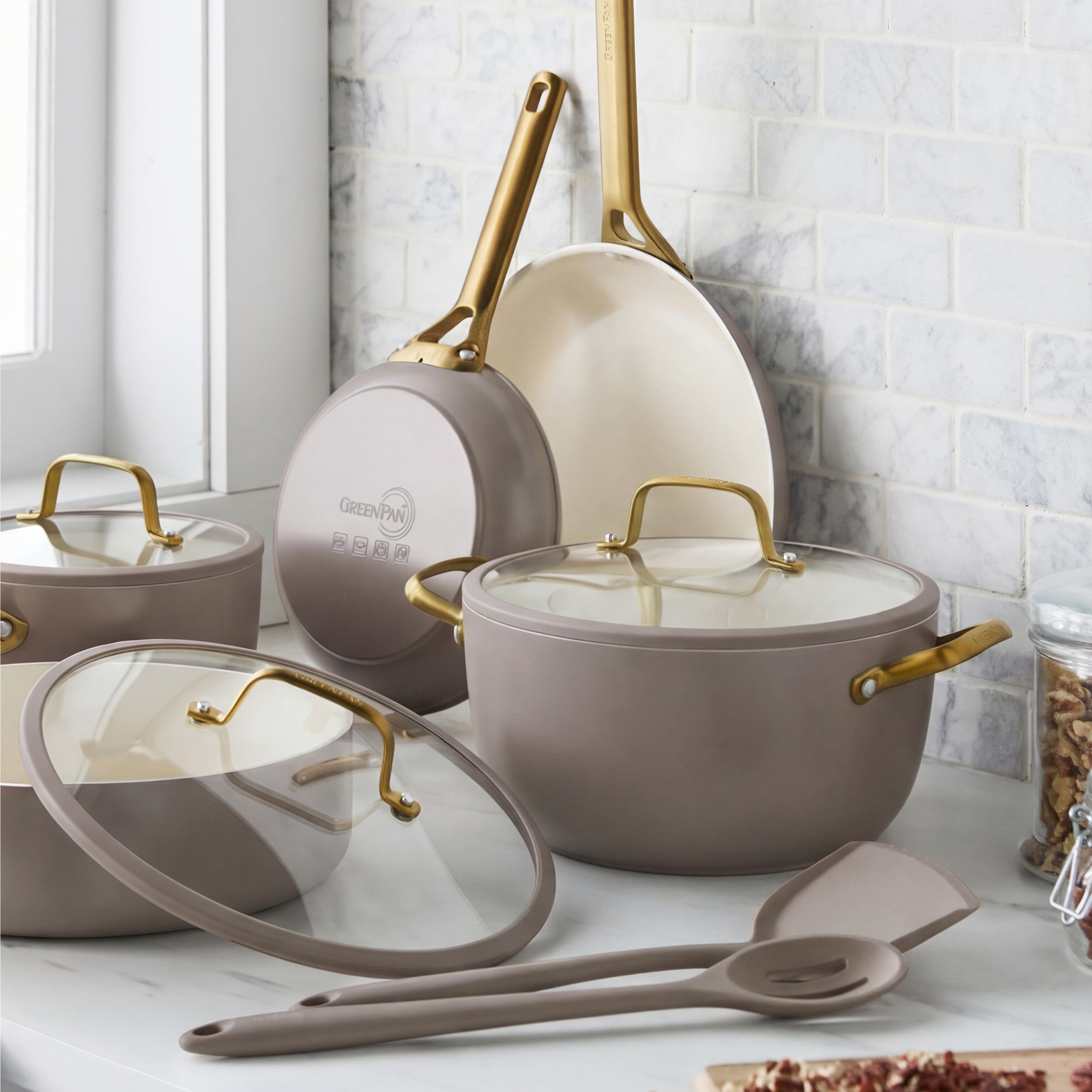 Latte-colored ceramic nonstick 10-piece cookware set on a marble counter beside a jar of nuts.