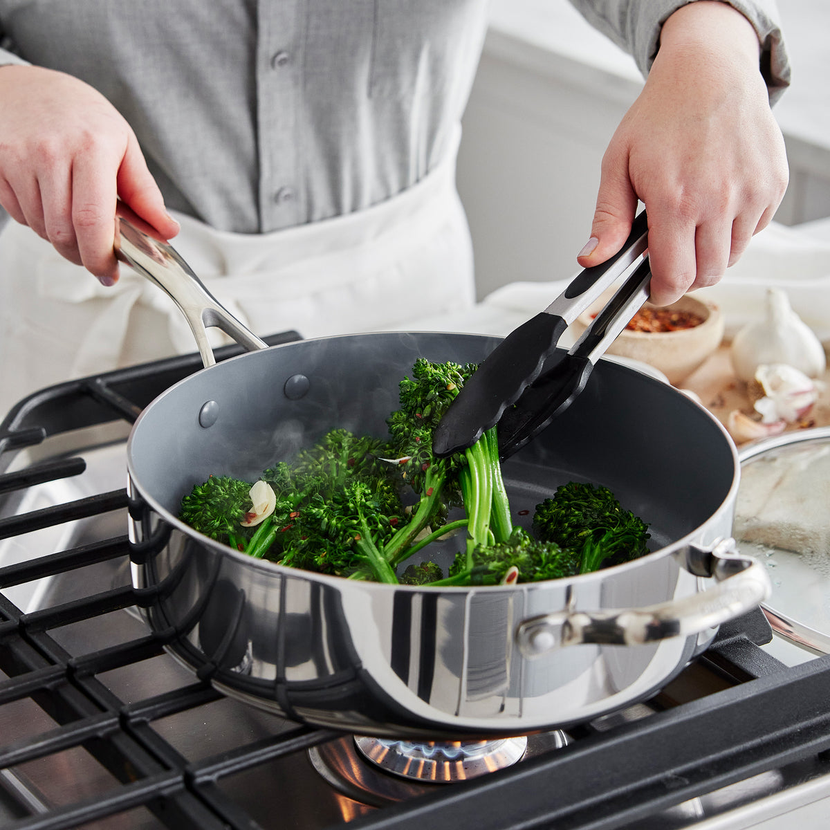 Broccolini being sautéed with black silicone tongs in a Valencia Pro stainless steel pan from an 18-piece cookware set on a gas stovetop.