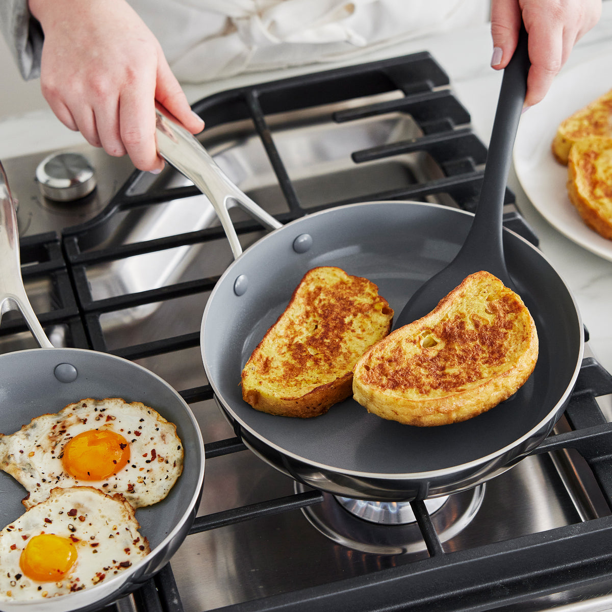 French toast being flipped with a spatula in a gray Valencia Pro 18-piece stainless steel pan next to a stainless steel pan.