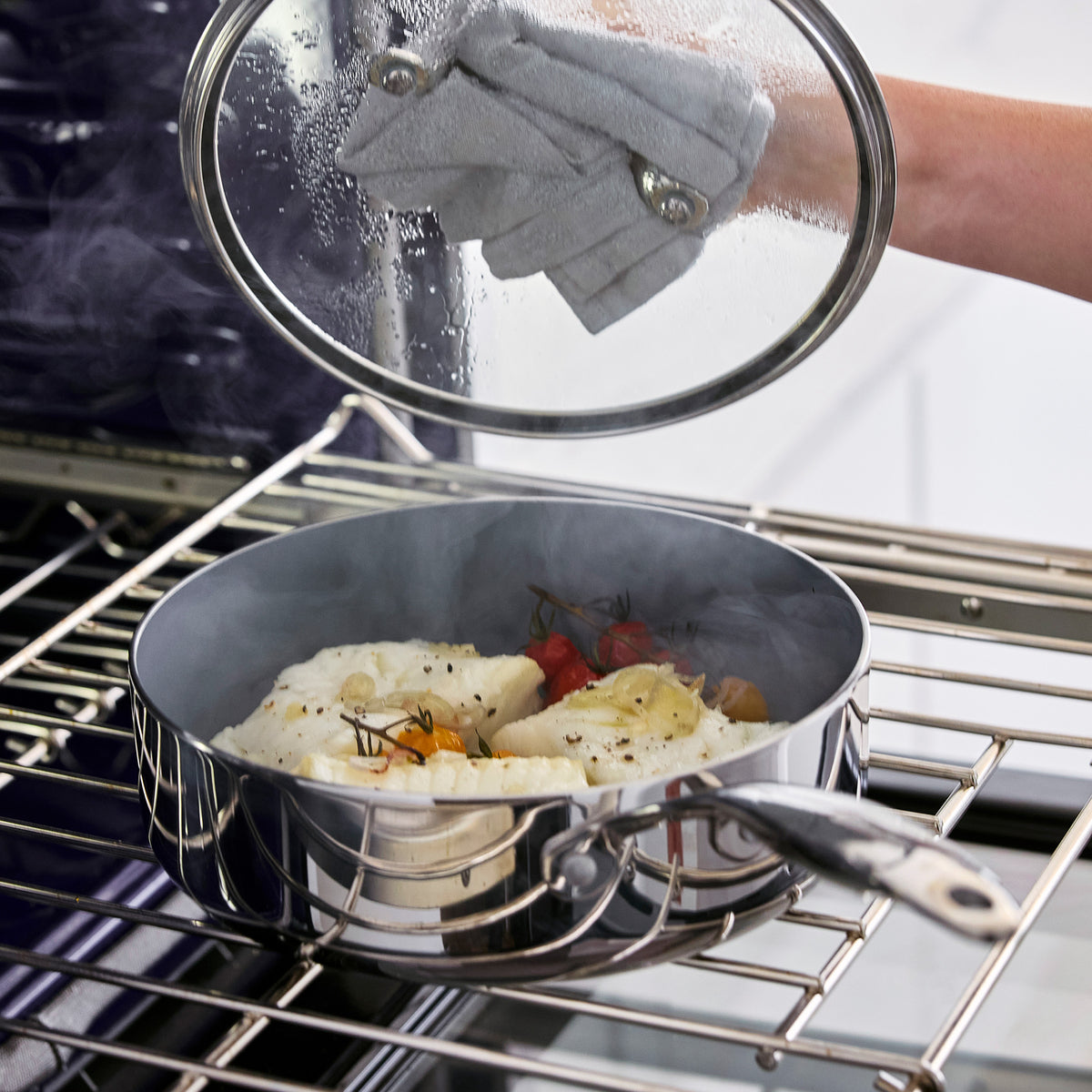 Hand in an oven mitt lifting the lid of a stainless steel Valencia Pro 18-piece cookware pan filled with fish and tomatoes.