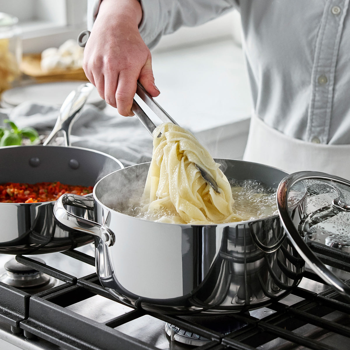Valencia Pro 18-piece stainless steel cookware set featuring a stockpot with steaming pasta and a ceramic saucepan.