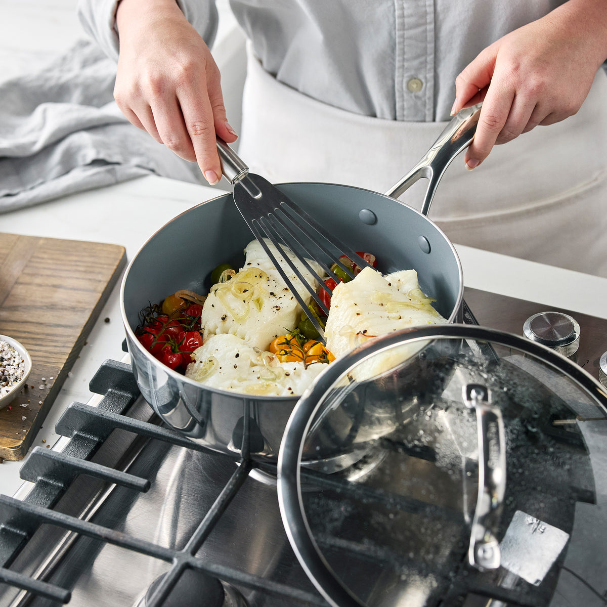 White fish and tomatoes being flipped with a spatula in a Valencia Pro stainless steel 18-piece cookware skillet on a stovetop.