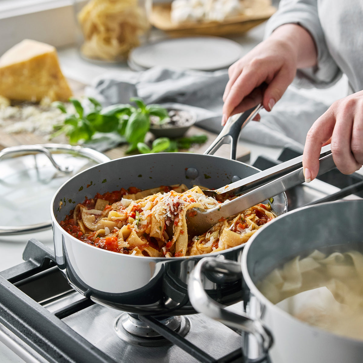 Metal tongs stirring pasta with tomato sauce in a stainless steel Valencia Pro 18-piece cookware skillet on a stovetop.