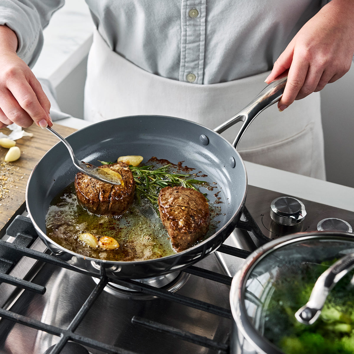 Gray Valencia Pro Stainless Steel 18-piece cookware pan on a gas stovetop with steaks searing alongside garlic and herbs.