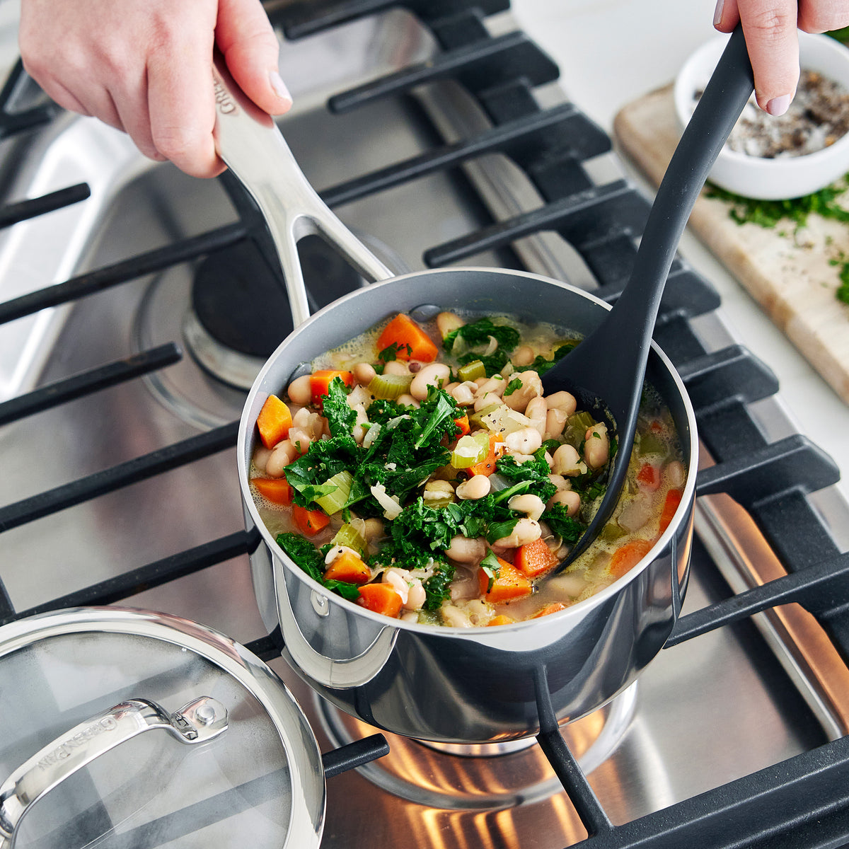 Valencia Pro stainless steel saucepan from the 18-piece set filled with bean and kale soup on a gas stovetop.