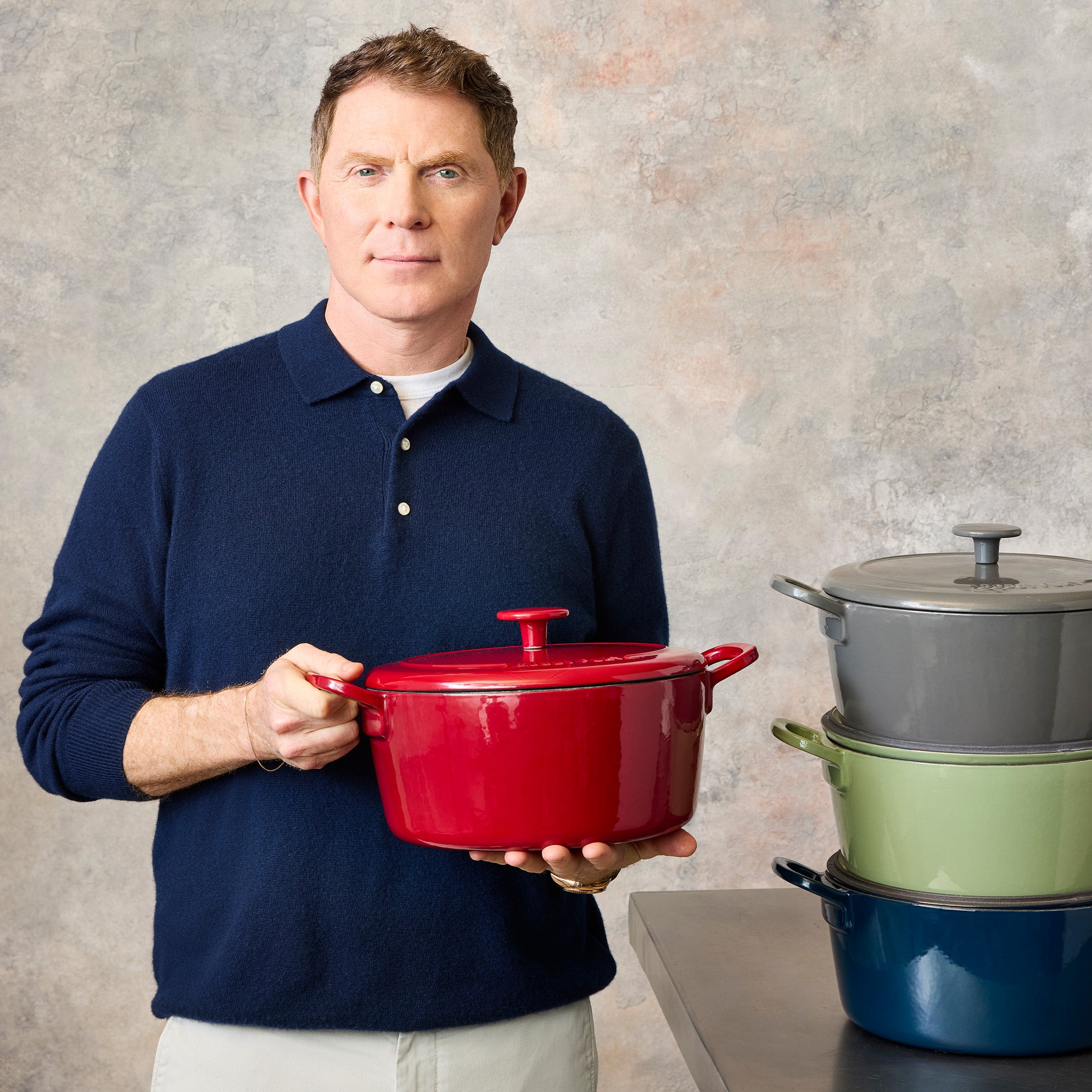 Man in navy shirt holding red Dutch oven with lid from Bobby Flay by GreenPan, next to a stack of colorful Dutch ovens.