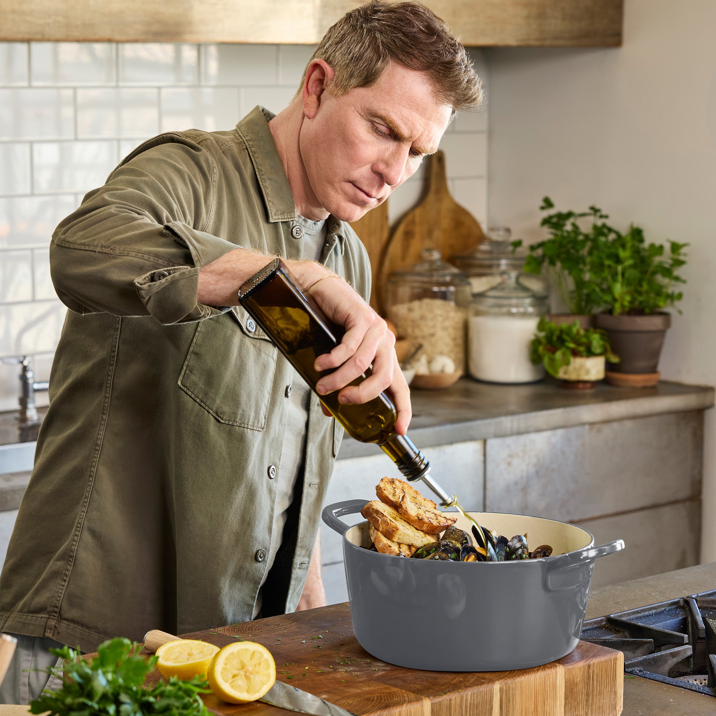 Man pours olive oil into a slate Dutch oven filled with seafood, bread, lemon slices, and herbs on a kitchen counter.