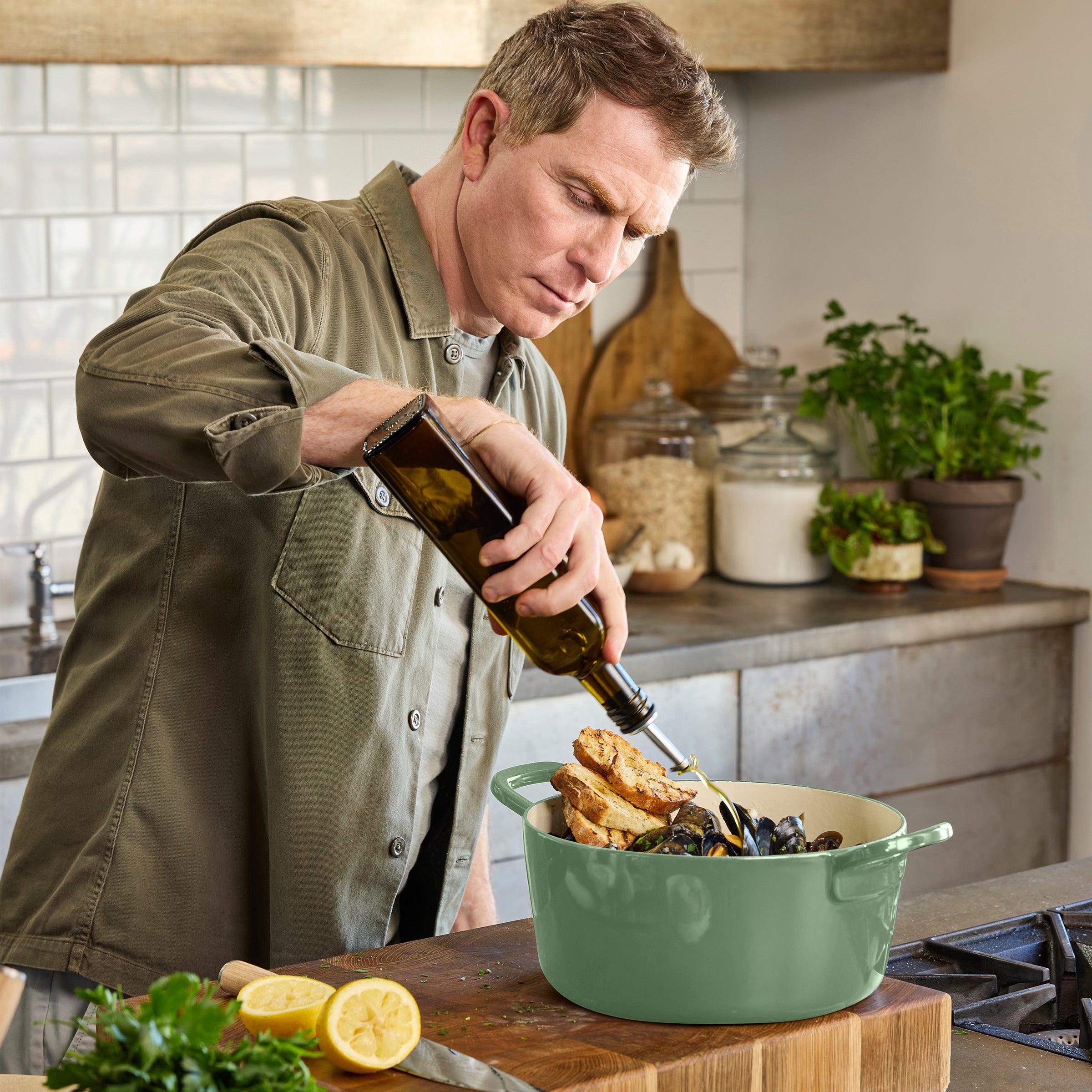 Mussels and bread in a sage green Dutch oven with olive oil being poured, herbs and lemons on the side.