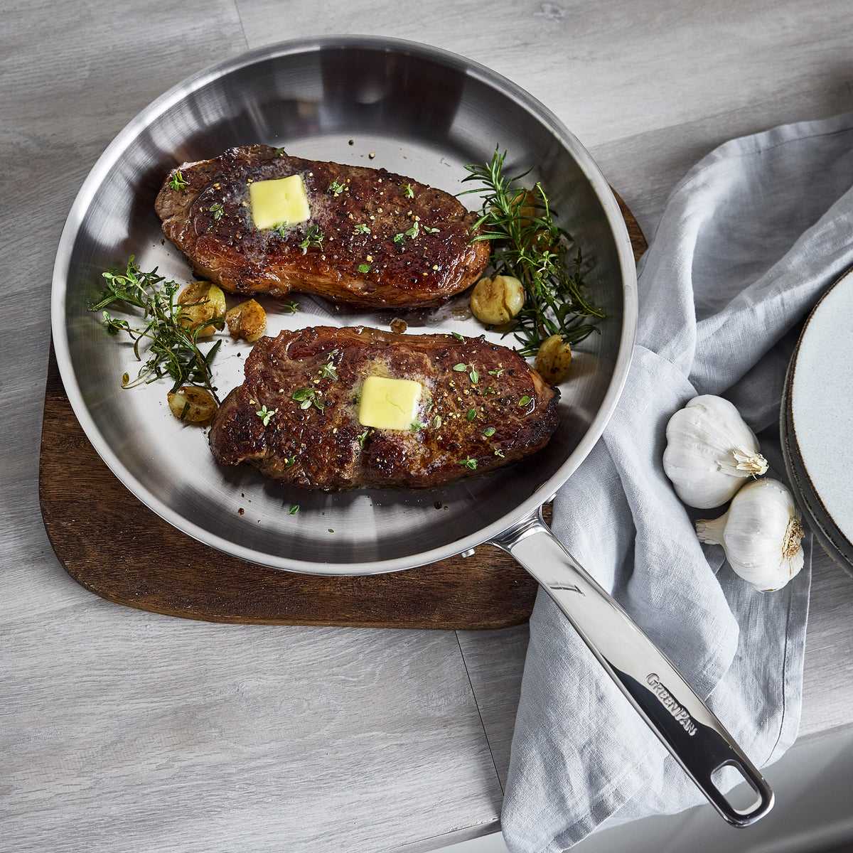 Two seared steaks topped with butter, herbs, and roasted garlic in a 12-inch Chatham Pro stainless steel frypan.