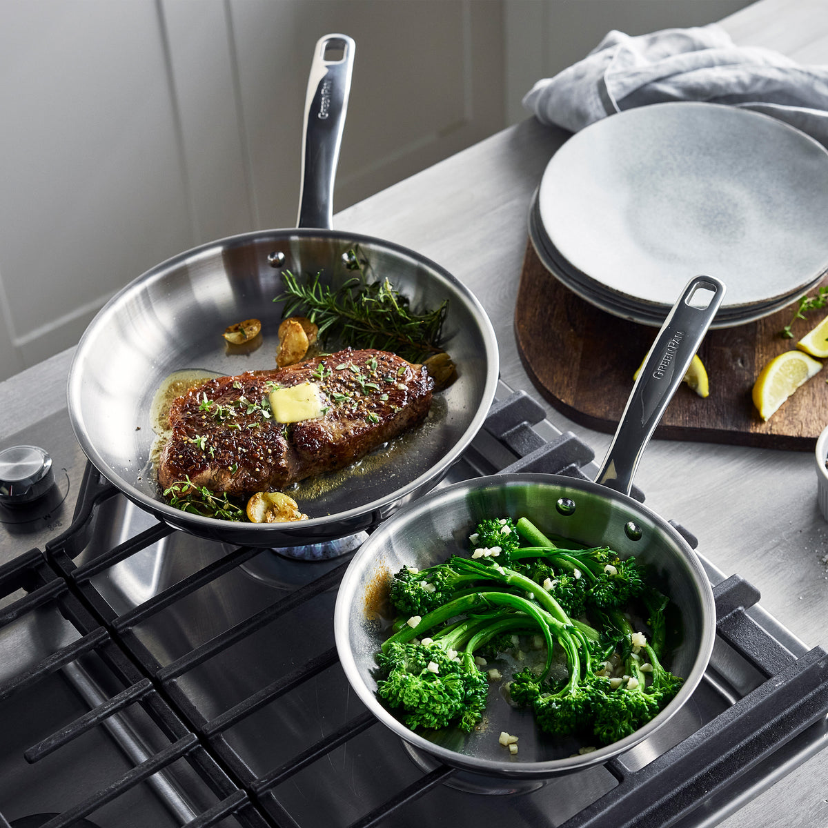 Two Chatham Pro 12-piece stainless steel pans on a stove: one searing steak with butter and herbs, the other sautéing broccolini.